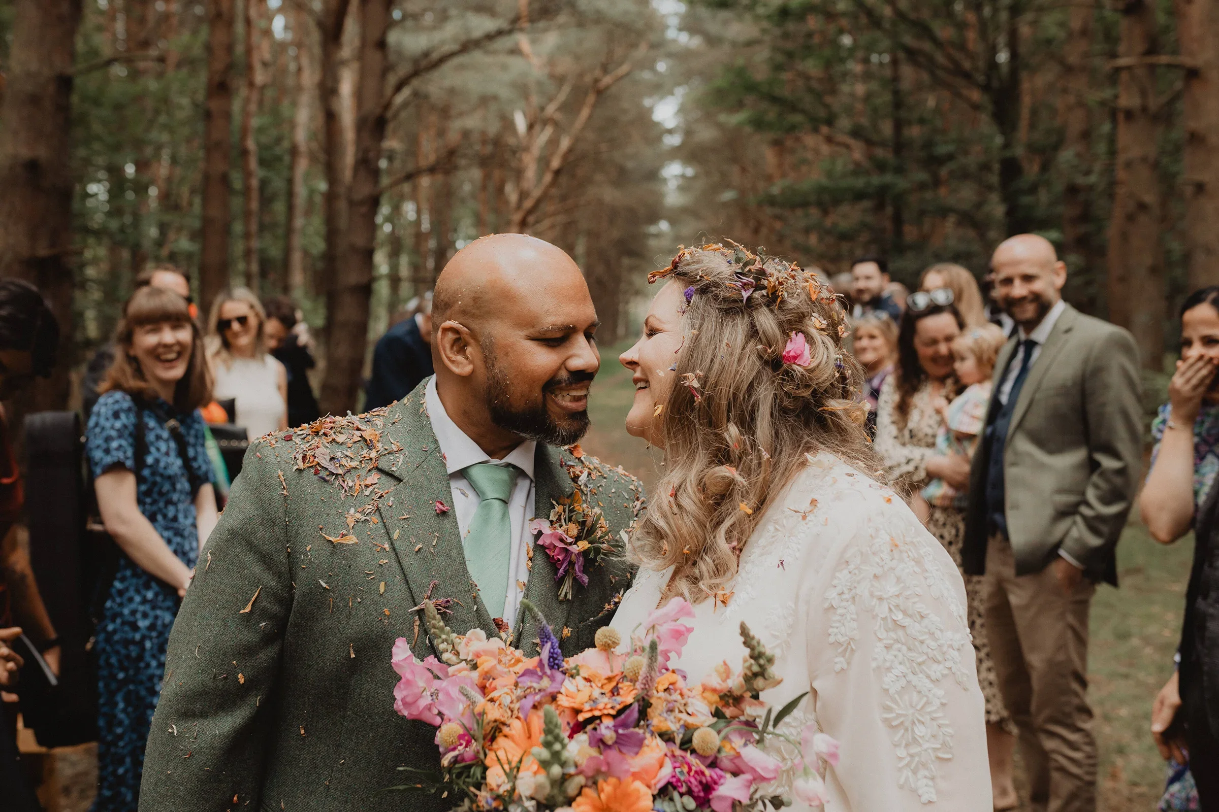 A couple gets married in a forest, surrounded by friends and family, with flower petals falling around them. The groom is wearing a green suit and tie, and the bride is holding a colorful bouquet. They are smiling and leaning close, with guests in th