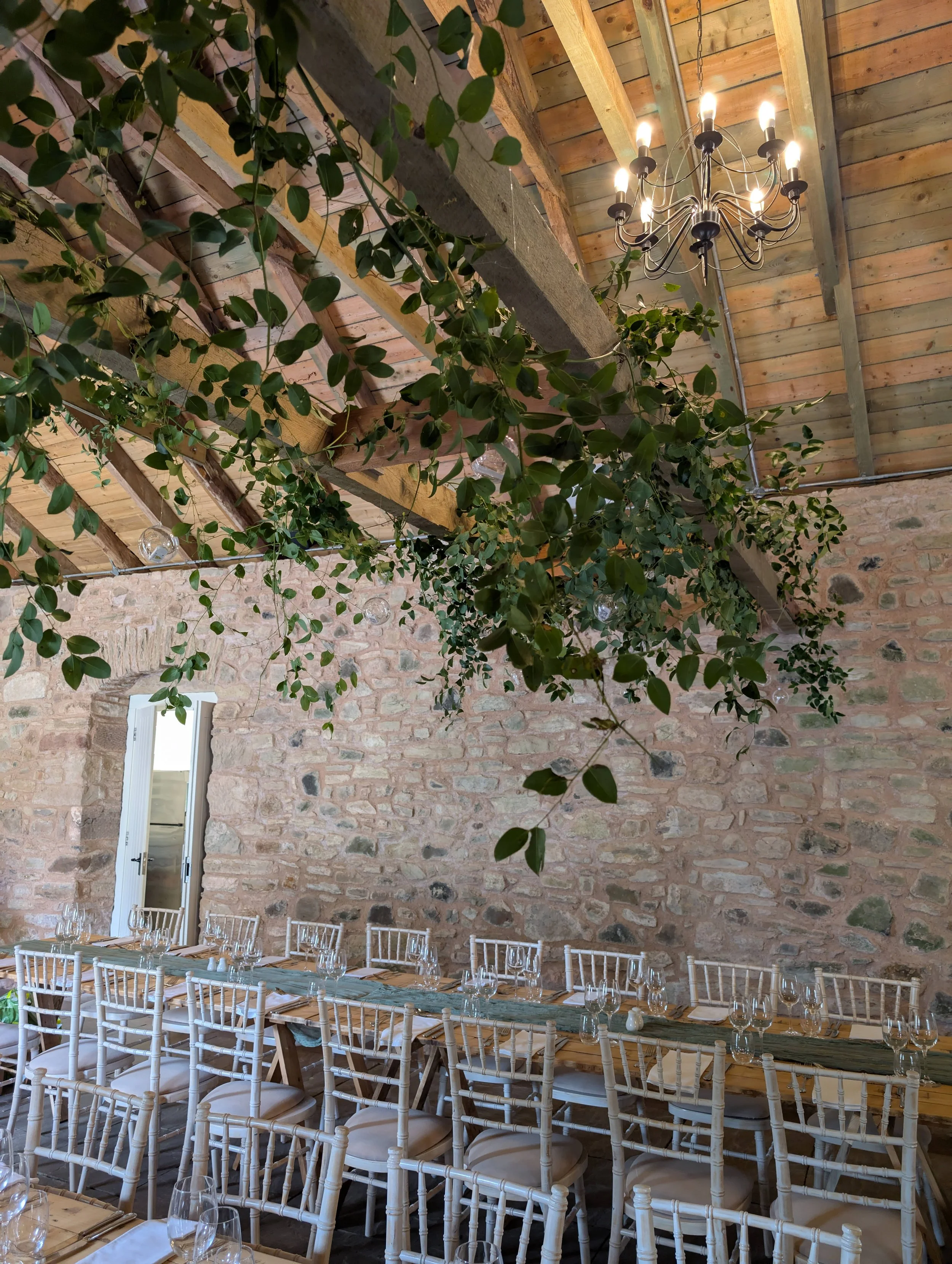 Indoor dining area with a long wooden table set with glassware and napkins, surrounded by white chairs with cushions, a stone wall, hanging greenery and string lights, and a chandelier on a wooden ceiling.