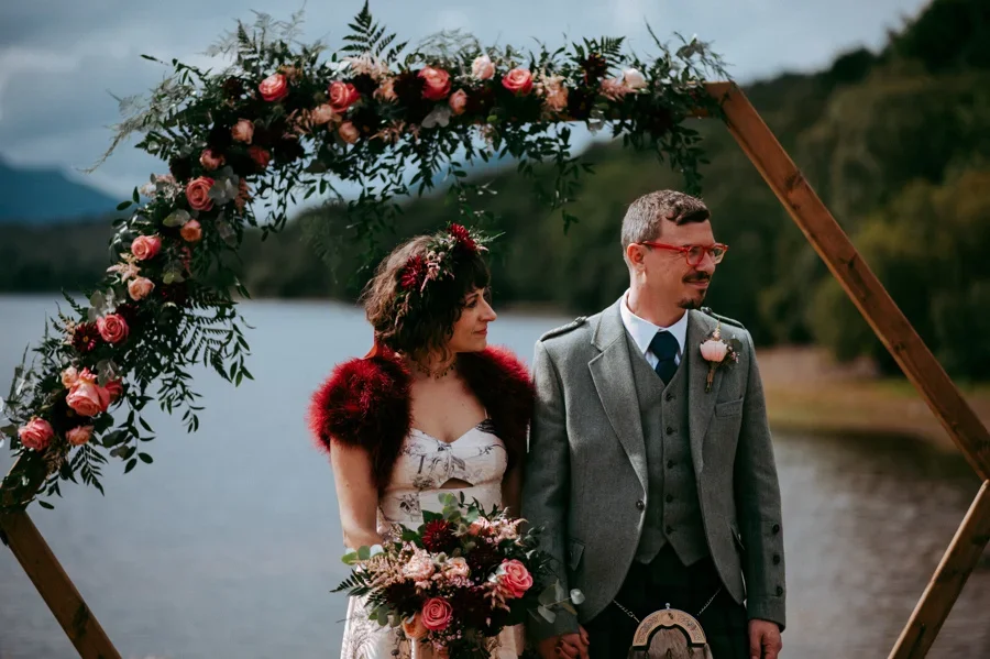 A couple at their outdoor wedding ceremony under a flower-decorated wooden arch near a lake, with mountains and trees in the background. The bride wears a floral dress with a red shawl and a flower crown, holding a bouquet. The groom wears a gray sui