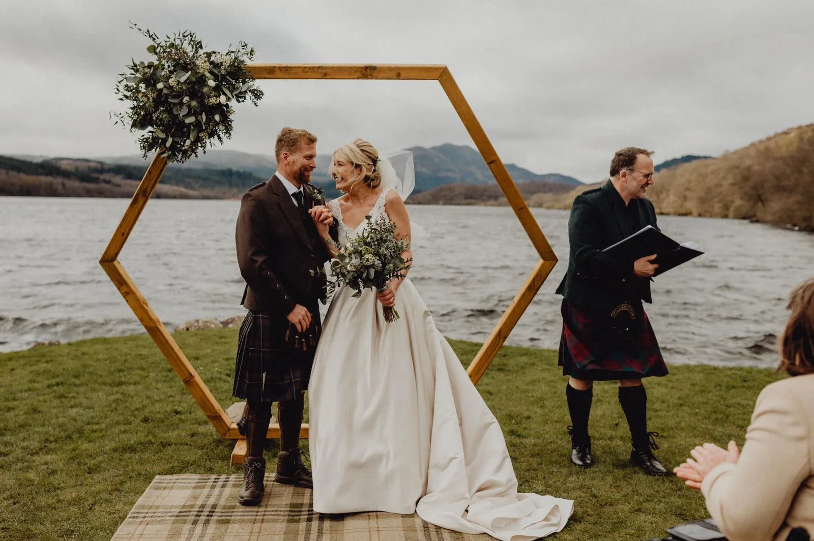 A bride and groom exchanging vows outdoors by a lake, with a hexagonal wooden arch decorated with greenery and white flowers, officiant holding a book, and a guest clapping in the foreground.