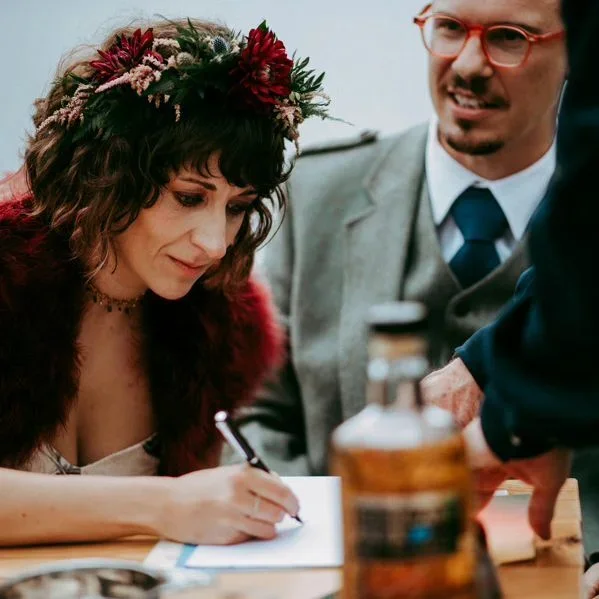 A woman with a floral crown signs a document at a table, while a man in glasses and a suit observes.