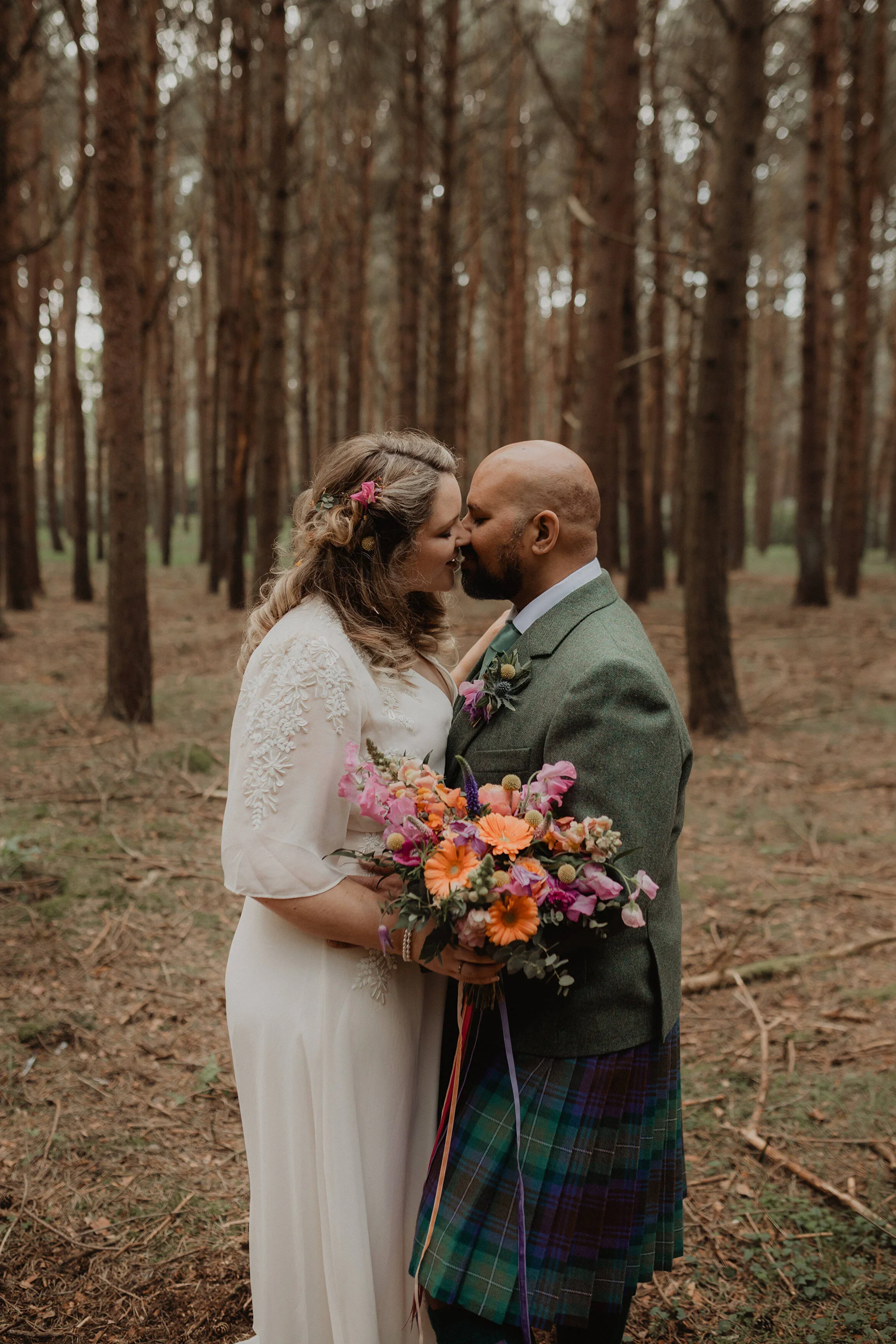 A couple kissing in a forest, the woman holding a colorful bouquet, both dressed in wedding attire.