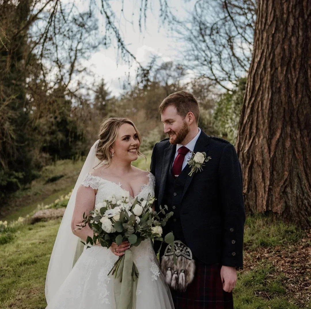 A bride and groom in wedding attire standing outdoors. The bride is holding a bouquet of white flowers and greenery. They are smiling at each other near a large tree in a natural, wooded setting.