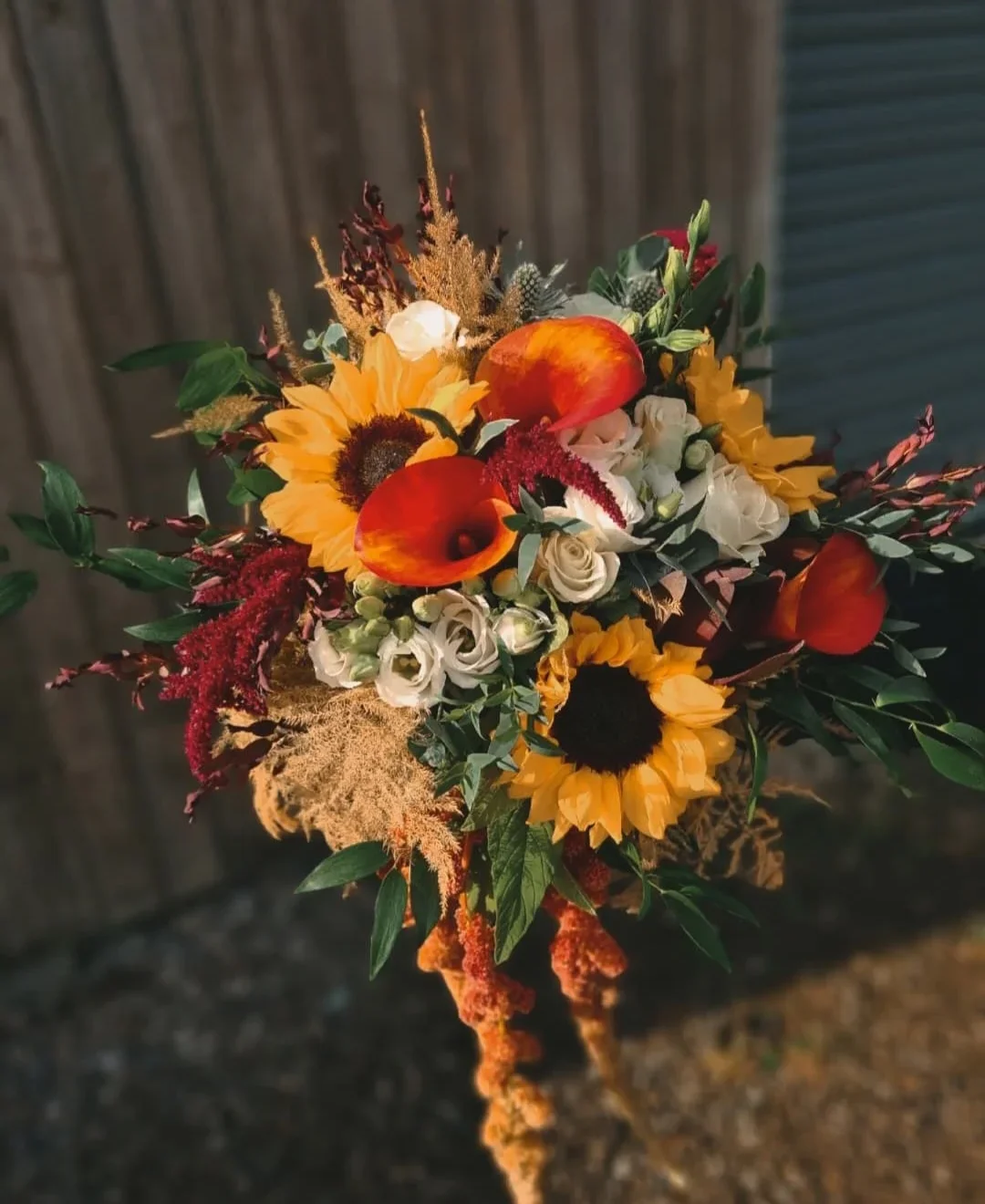 A vibrant bouquet of sunflowers, white roses, red and orange calla lilies, and assorted greenery against a wooden background.