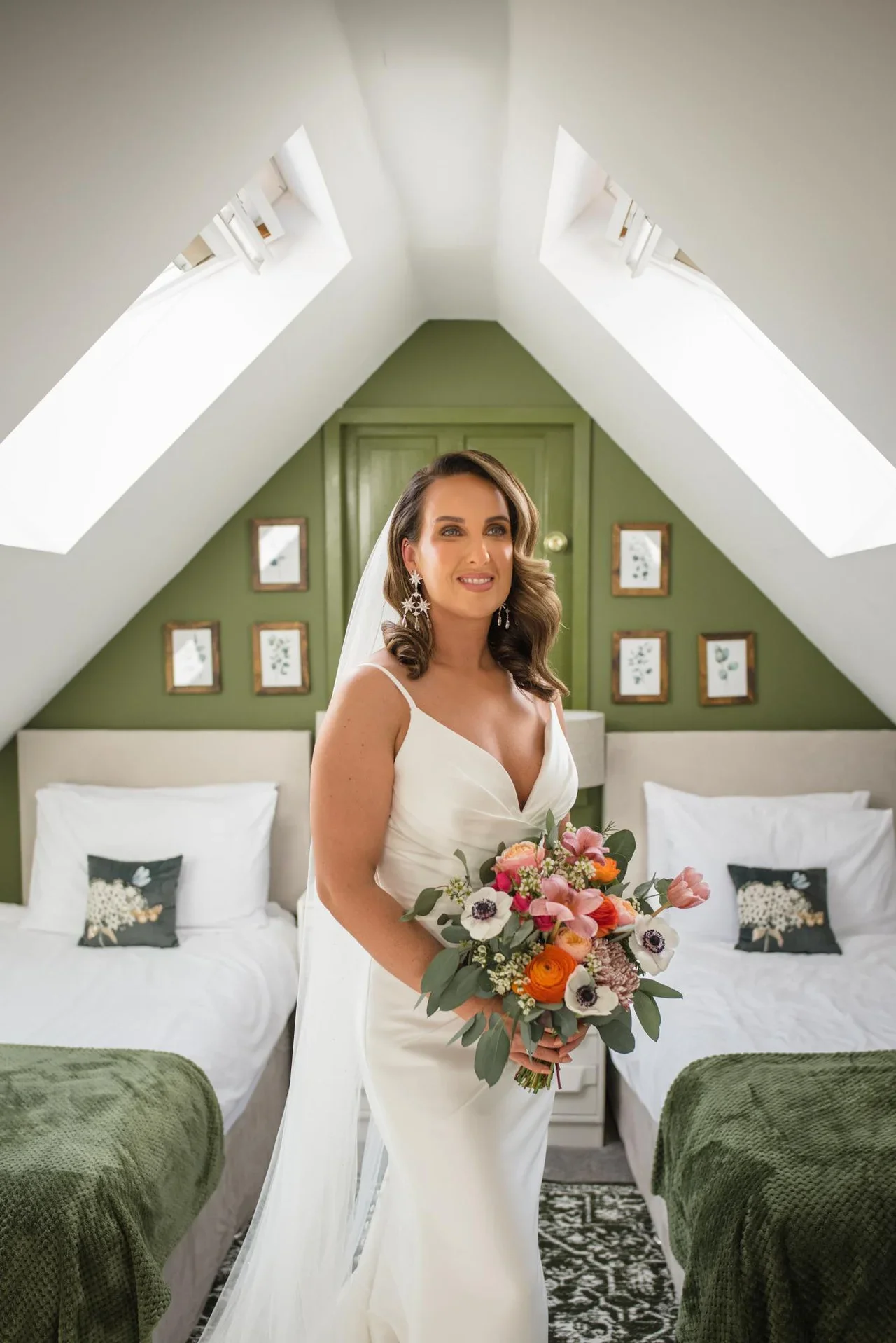 A bride in a white wedding dress holding a colorful bouquet of flowers stands in a cozy attic room with green walls, framed botanical artwork, and twin beds with white linens.