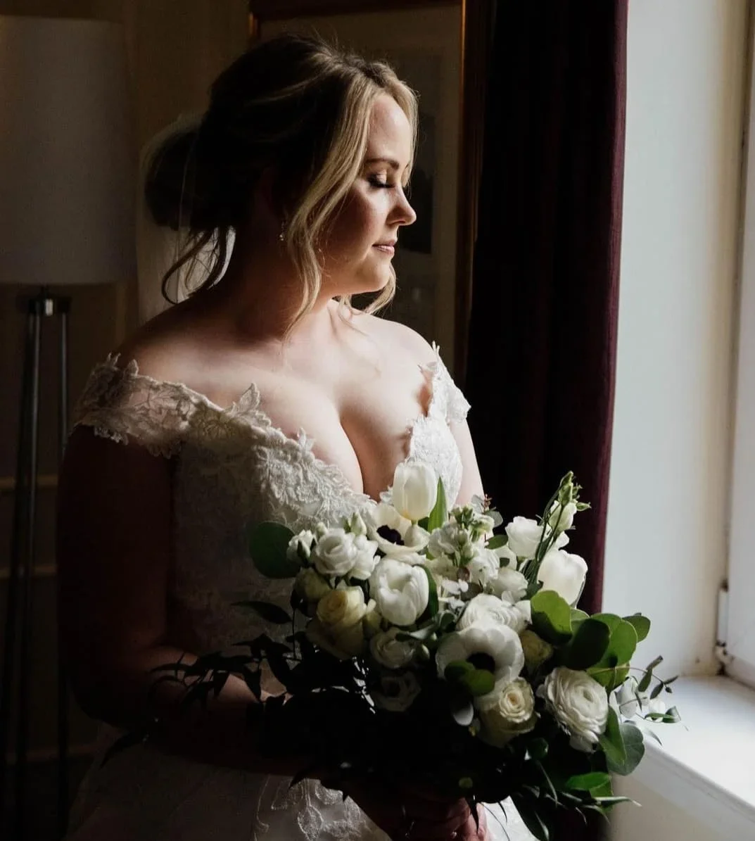A bride stands by a window holding a bouquet of white flowers, dressed in a lace wedding gown, with soft lighting highlighting her profile and the flowers.