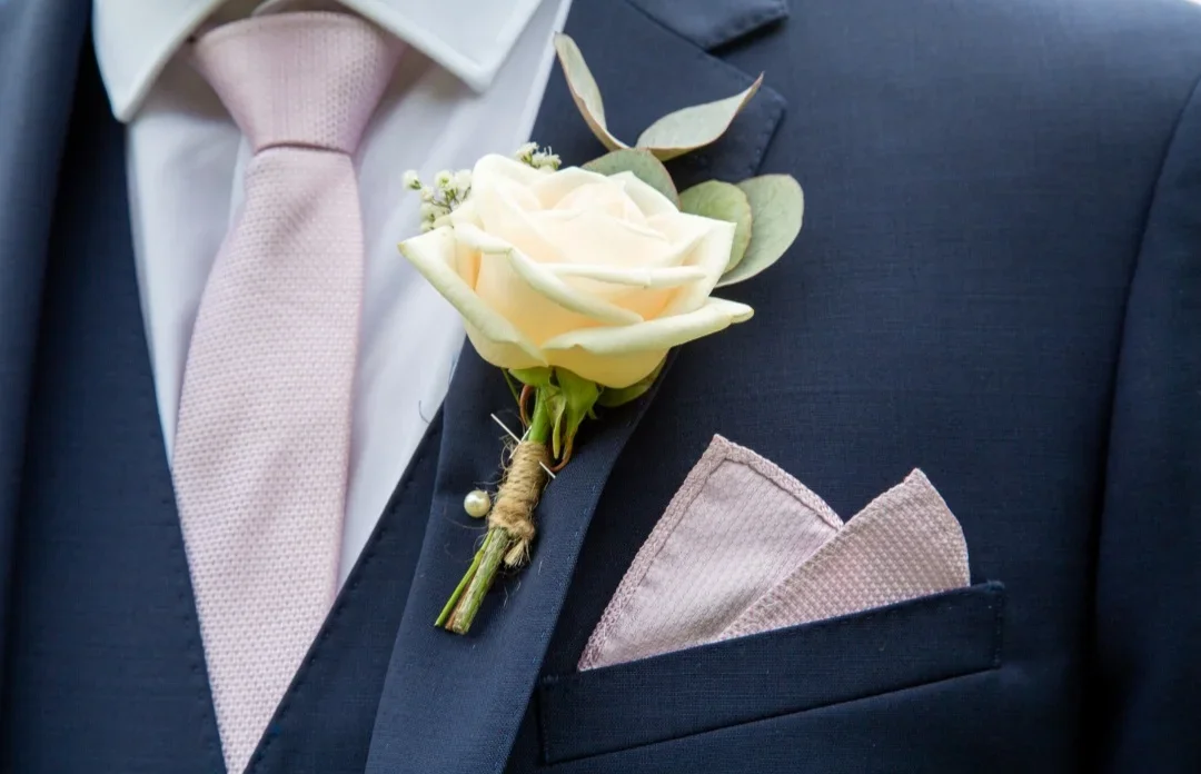 Close-up of a man in a navy suit with a pink tie, pink pocket square, and a white rose boutonniere with greenery on the lapel.