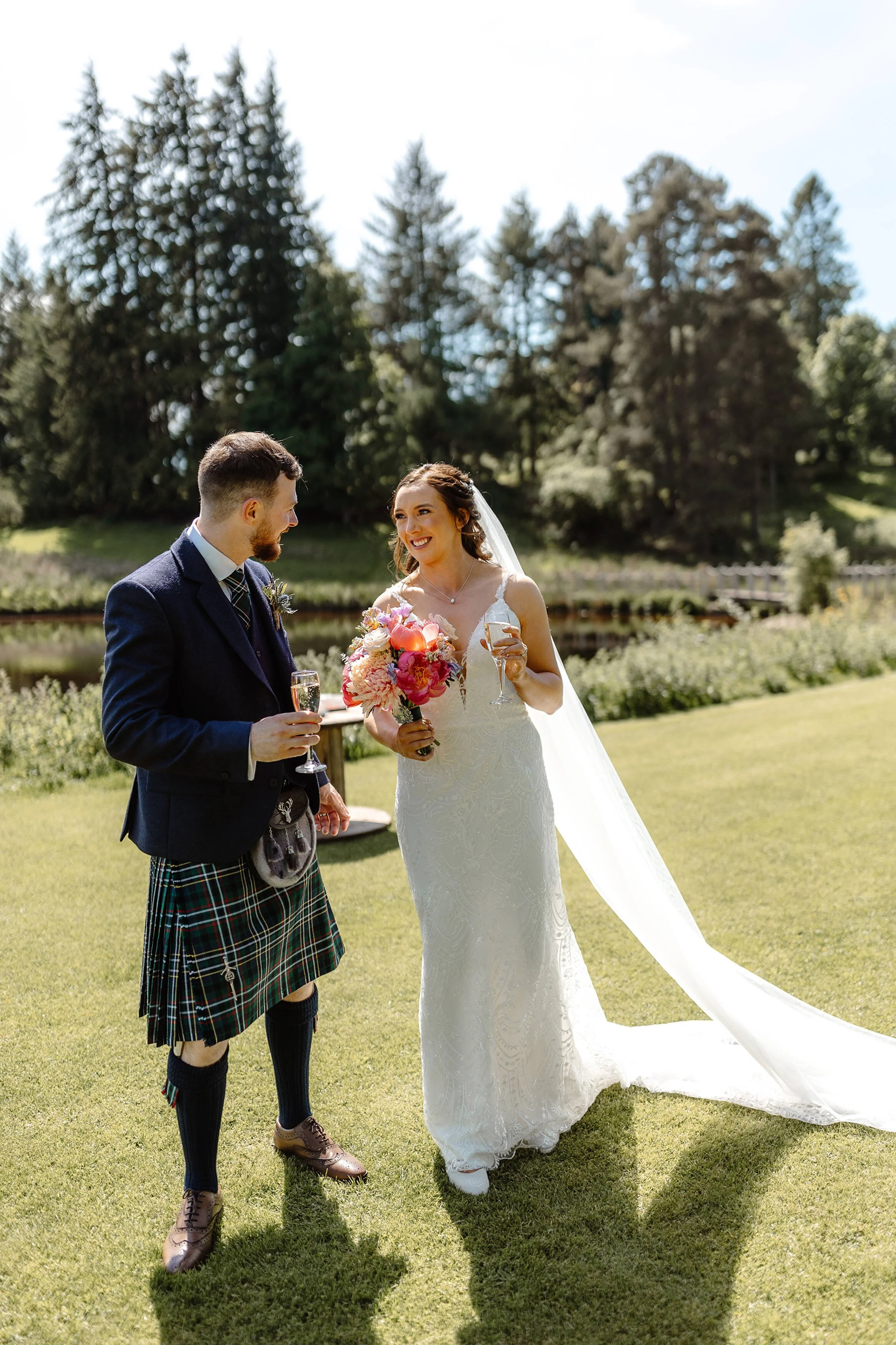 A bride and groom at an outdoor wedding celebration, holding glasses of champagne, standing on grass near a river, with trees in the background.