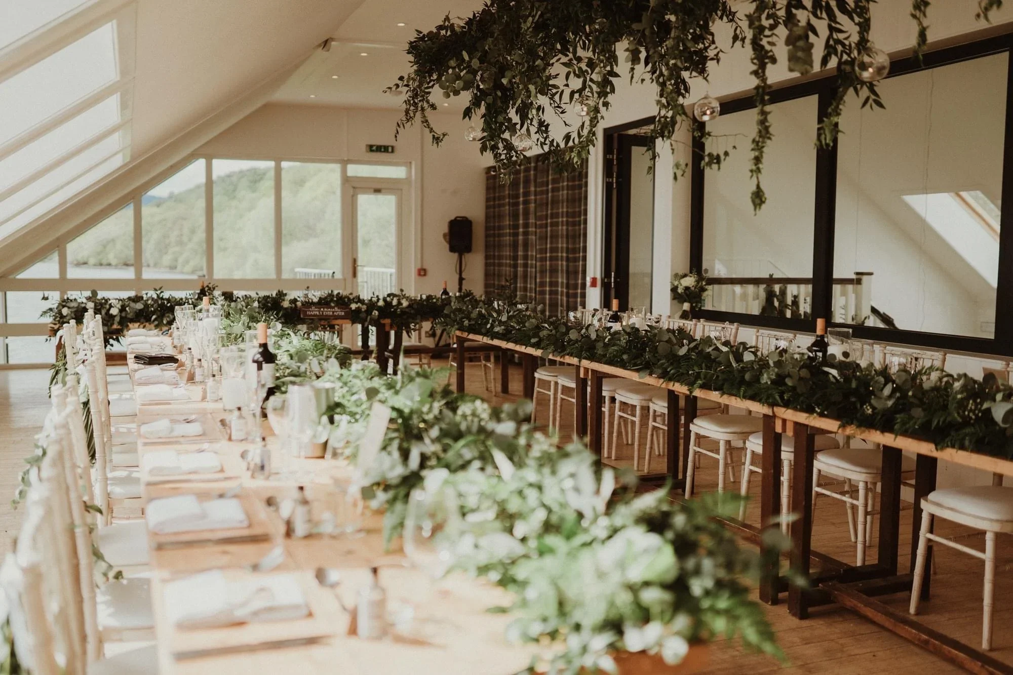 Elegant wedding reception setup with long wooden tables decorated with greenery garlands, white plates, silverware, wine bottles, and glasses. The room has large windows, a slanted ceiling with skylights, and a cozy ambiance with natural light.