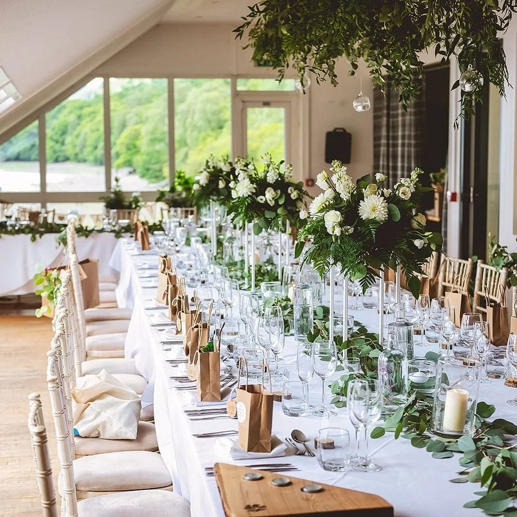 Elegant wedding reception table decorated with white flowers, greenery, glassware, and small paper gift bags, set in a room with large windows showing a green outdoor landscape.