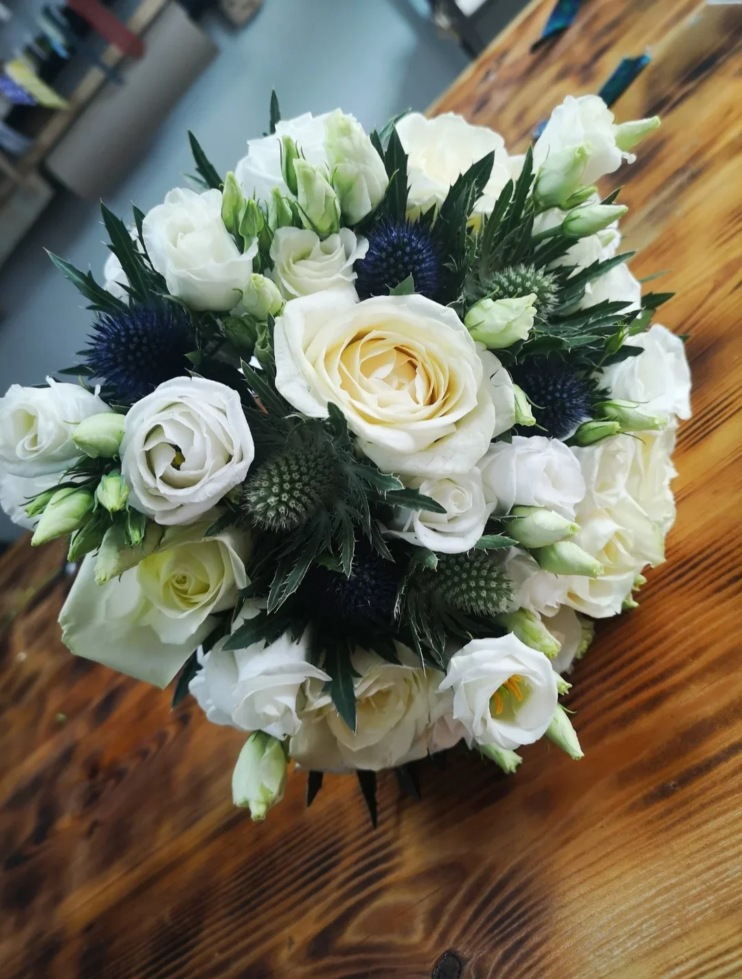 A bouquet of white roses, lisianthus, thistles, and greenery arranged in a round shape on a wooden table.