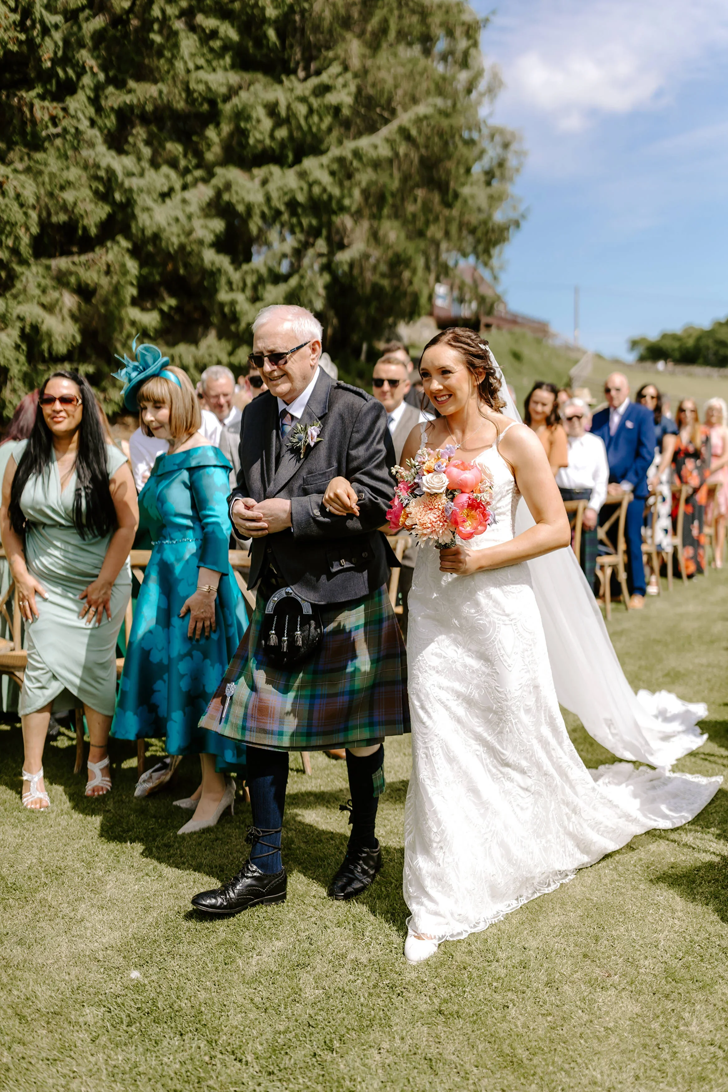 Bride walking down the aisle with an older man, likely her father, at an outdoor wedding on a sunny day with rows of seated guests and green trees in the background.