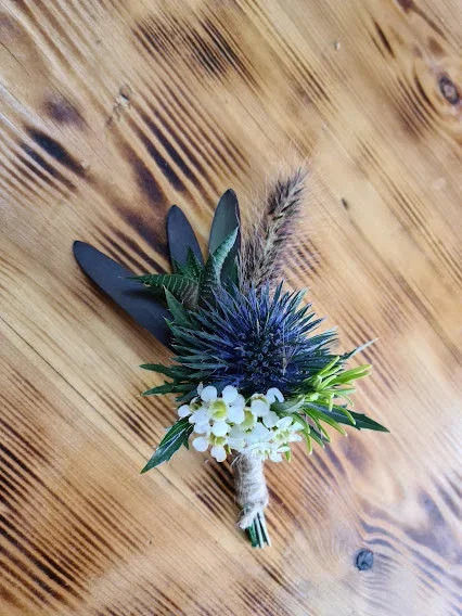 Boutonniere with purple thistle, white flowers, and green foliage, tied with twine, placed on a wooden surface.