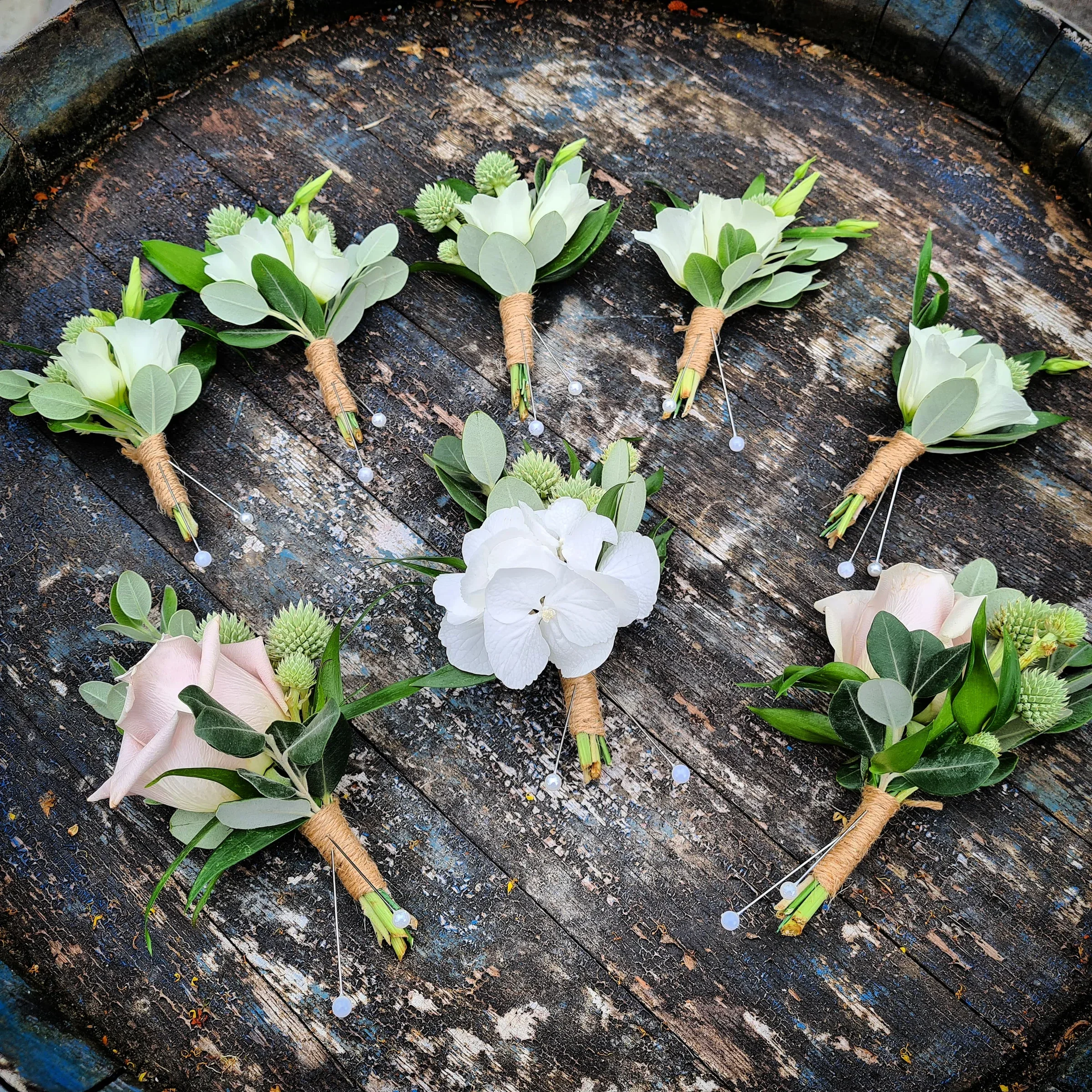 A heart-shaped arrangement of boutonnières and corsages with white and blush pink flowers, green leaves, wrapped in twine, placed on a rustic wooden surface.