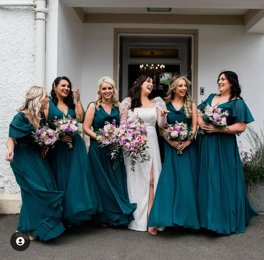 A bride and five bridesmaids standing outside a building, all laughing and holding bouquets of pink and purple flowers. The bride is wearing a white dress with a slit, while the bridesmaids are in teal dresses.