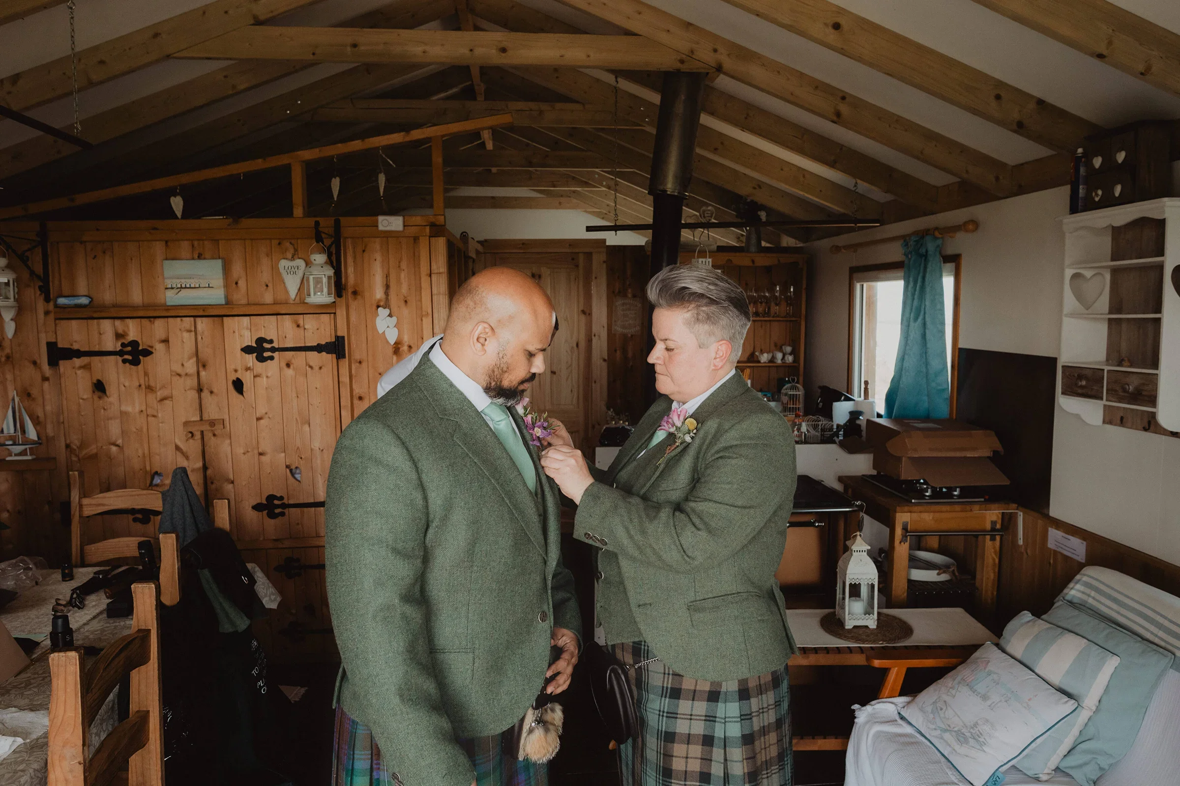 Two men in green blazers and tartan skirts engaging in a heartfelt moment inside a cozy wooden room, possibly during a wedding or special occasion.