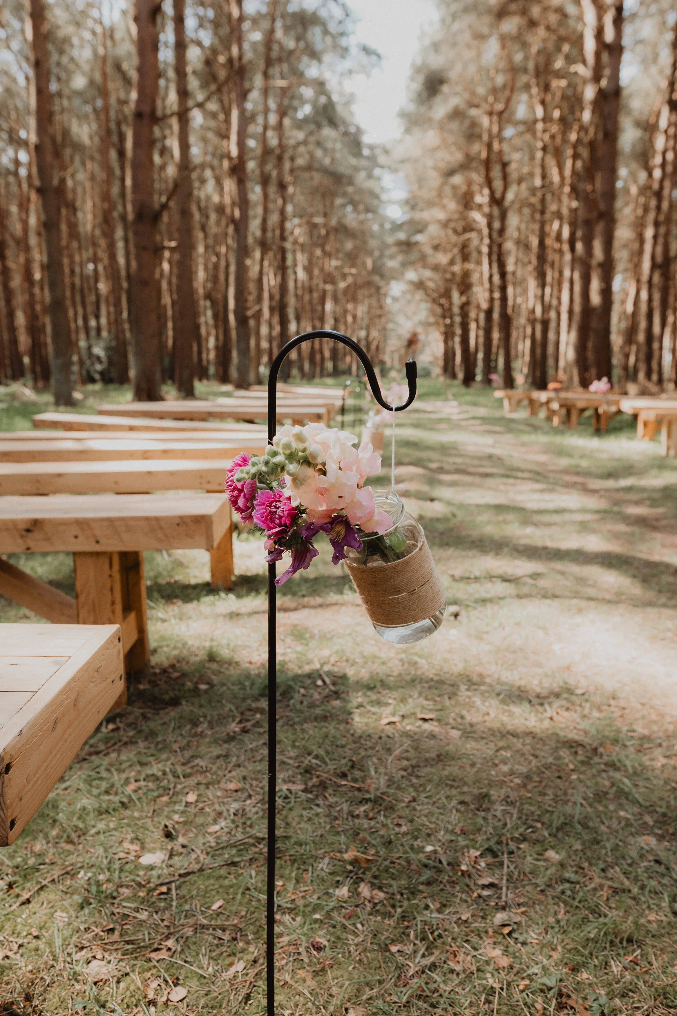 A wedding or outdoor event setup in a forest with arranged wooden benches, and a decorative mason jar with flowers hanging from a black metal stand.