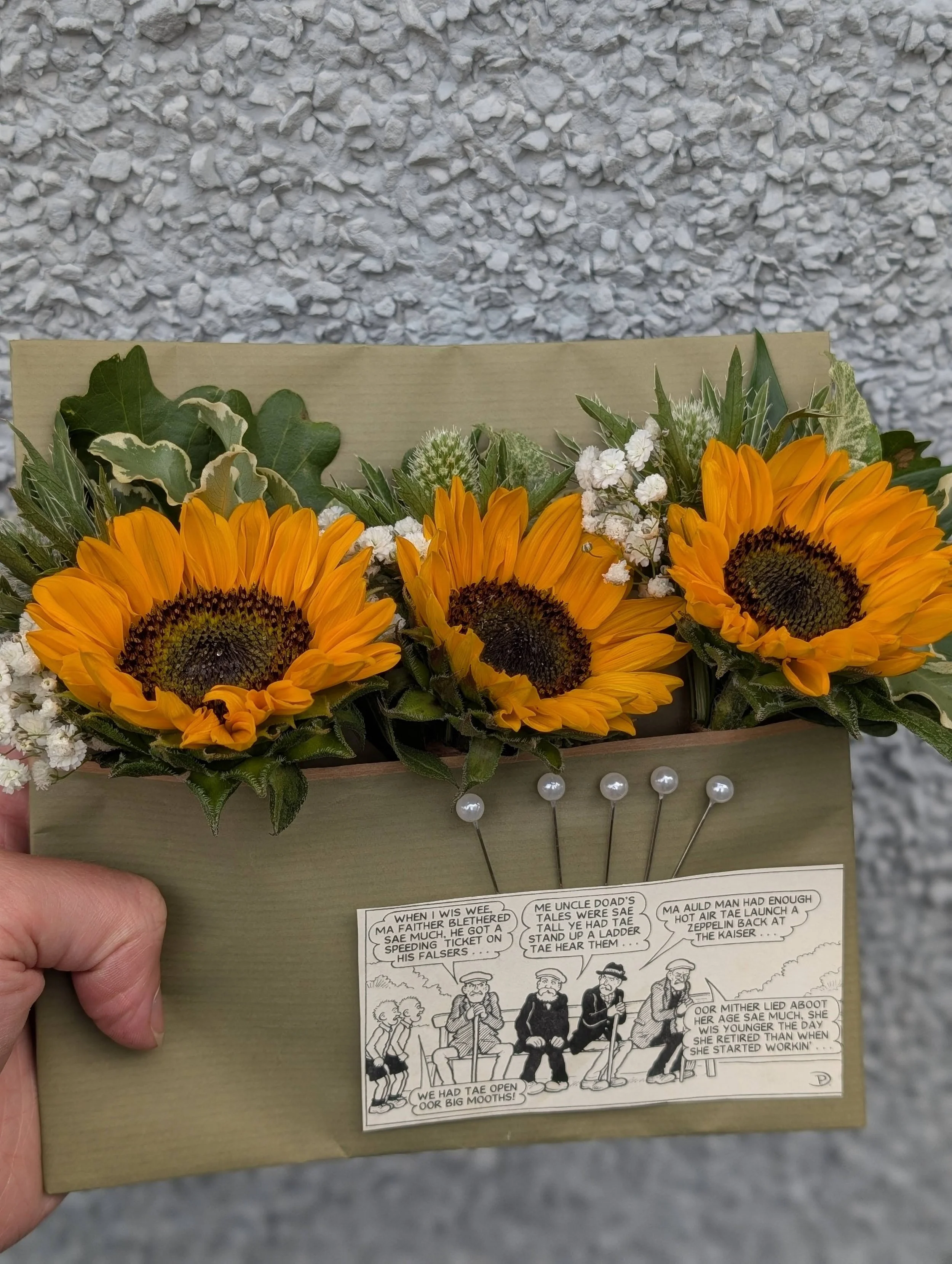 A hand holding a floral arrangement of three sunflowers, white baby's breath, and various green leaves, arranged in a cardboard gift box with a comic strip on the front side.