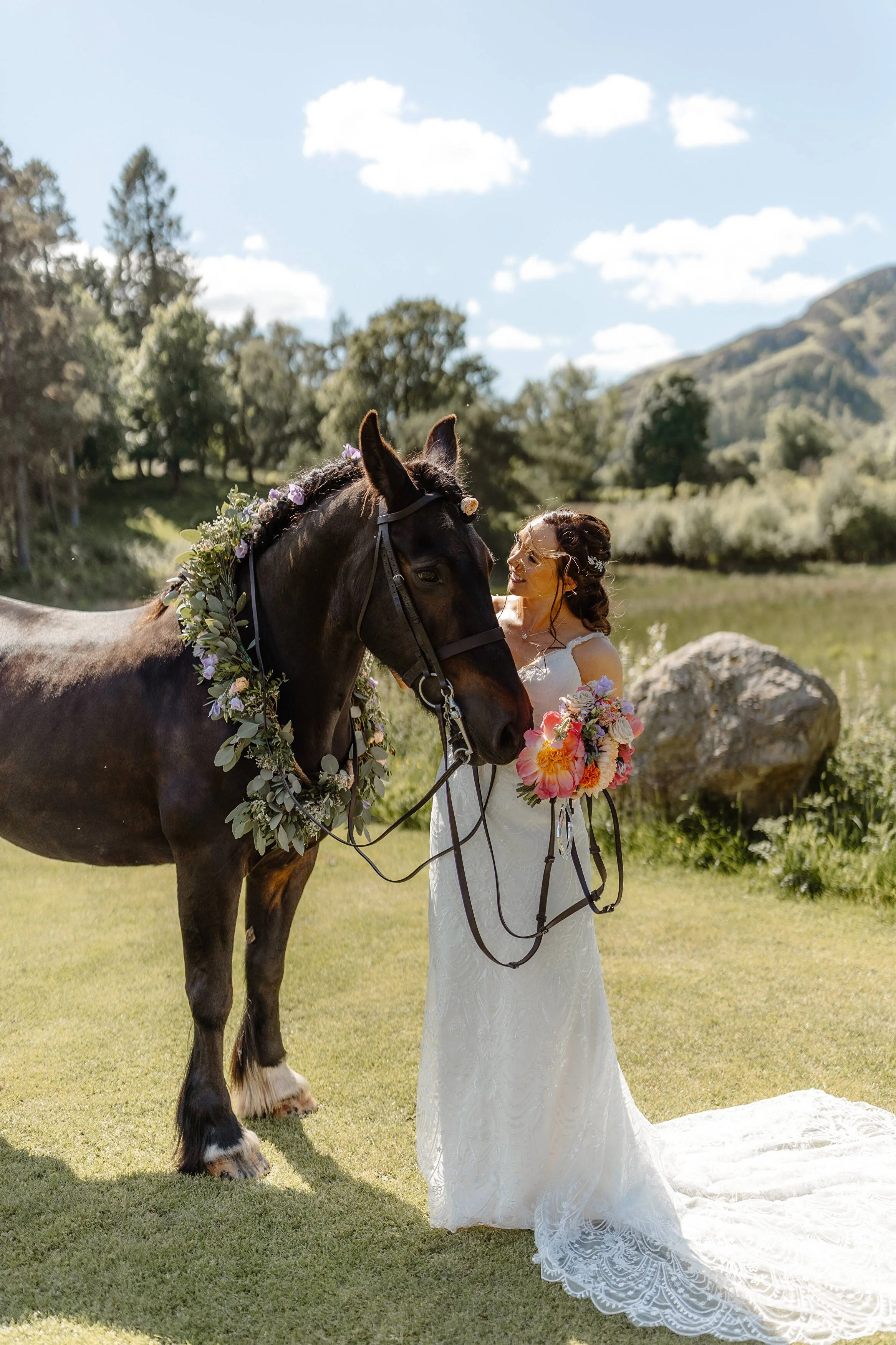 Bride in a white lace wedding dress holding a bouquet standing next to a decorated black horse in an outdoor natural setting.