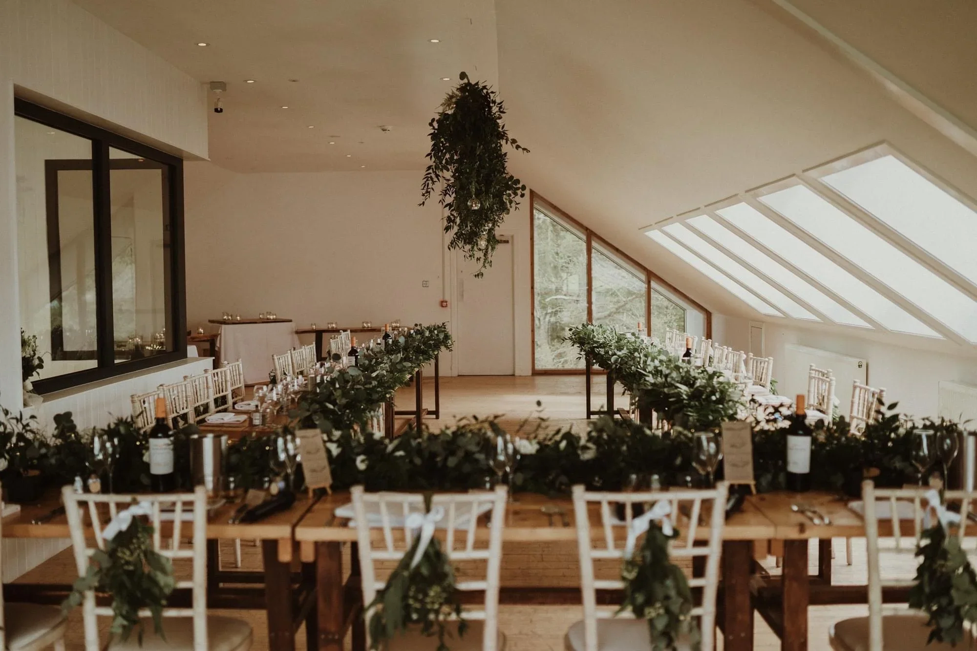 Elegant wedding reception set up in a bright, modern room with a sloped ceiling and large windows, featuring a long wooden table decorated with greenery and wine bottles, and white chairs with ribbons.