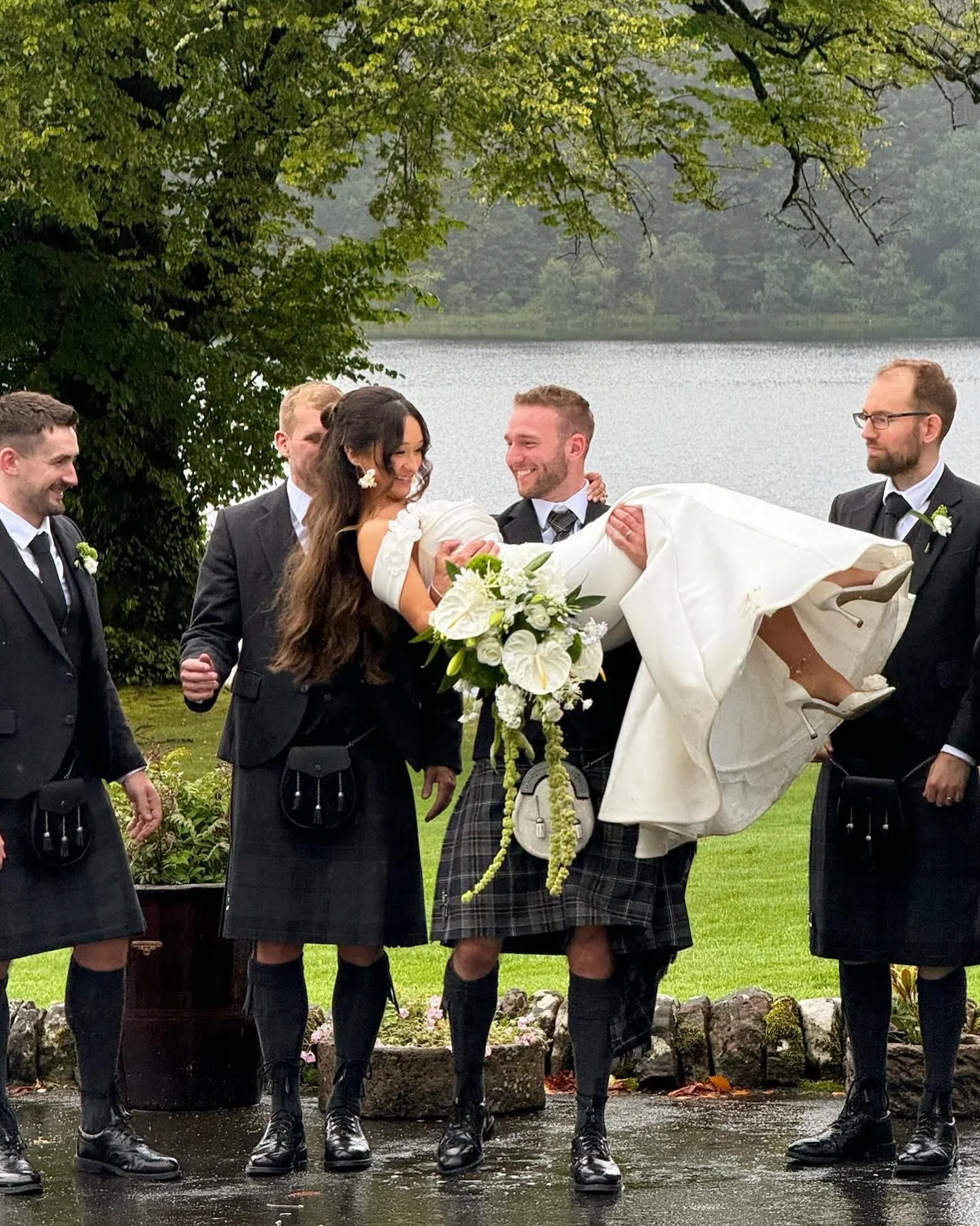 A bride being carried by a groom and surrounded by groomsmen and bridesmaids in Scottish kilts outdoor by a lake.
