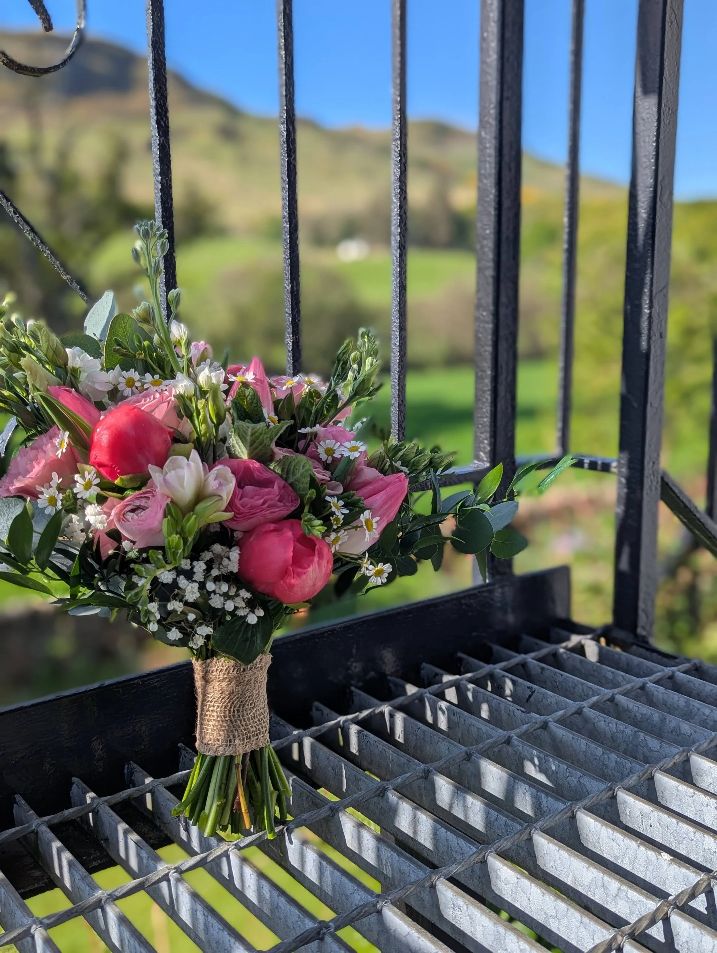 A bouquet of pink and white flowers tied with burlap ribbon resting on a metal grate railing, with a blurred green landscape and hills in the background.