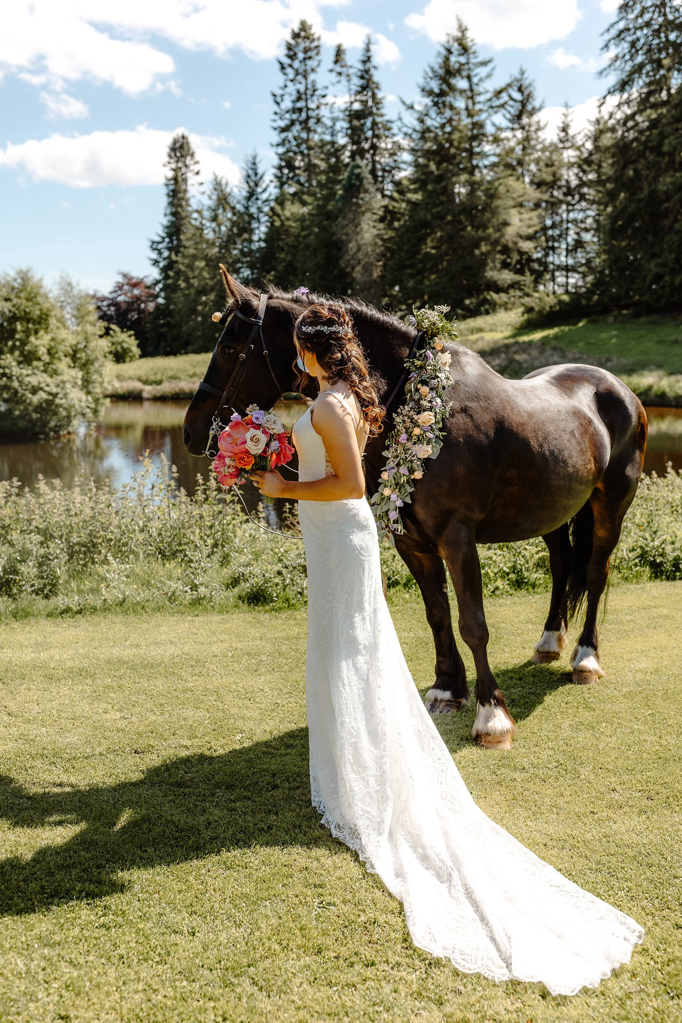 A woman in a white wedding dress holding a bouquet of pink and white flowers and standing next to a decorated black horse with floral garland, by a pond and trees under a blue sky.