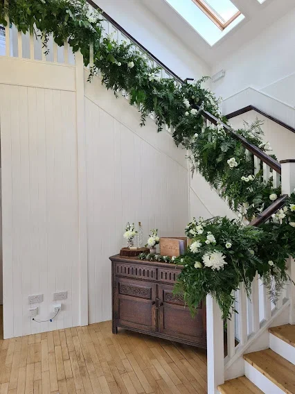 Decorated staircase with green foliage and white flowers, wooden sideboard underneath, and skylight above in a bright room.