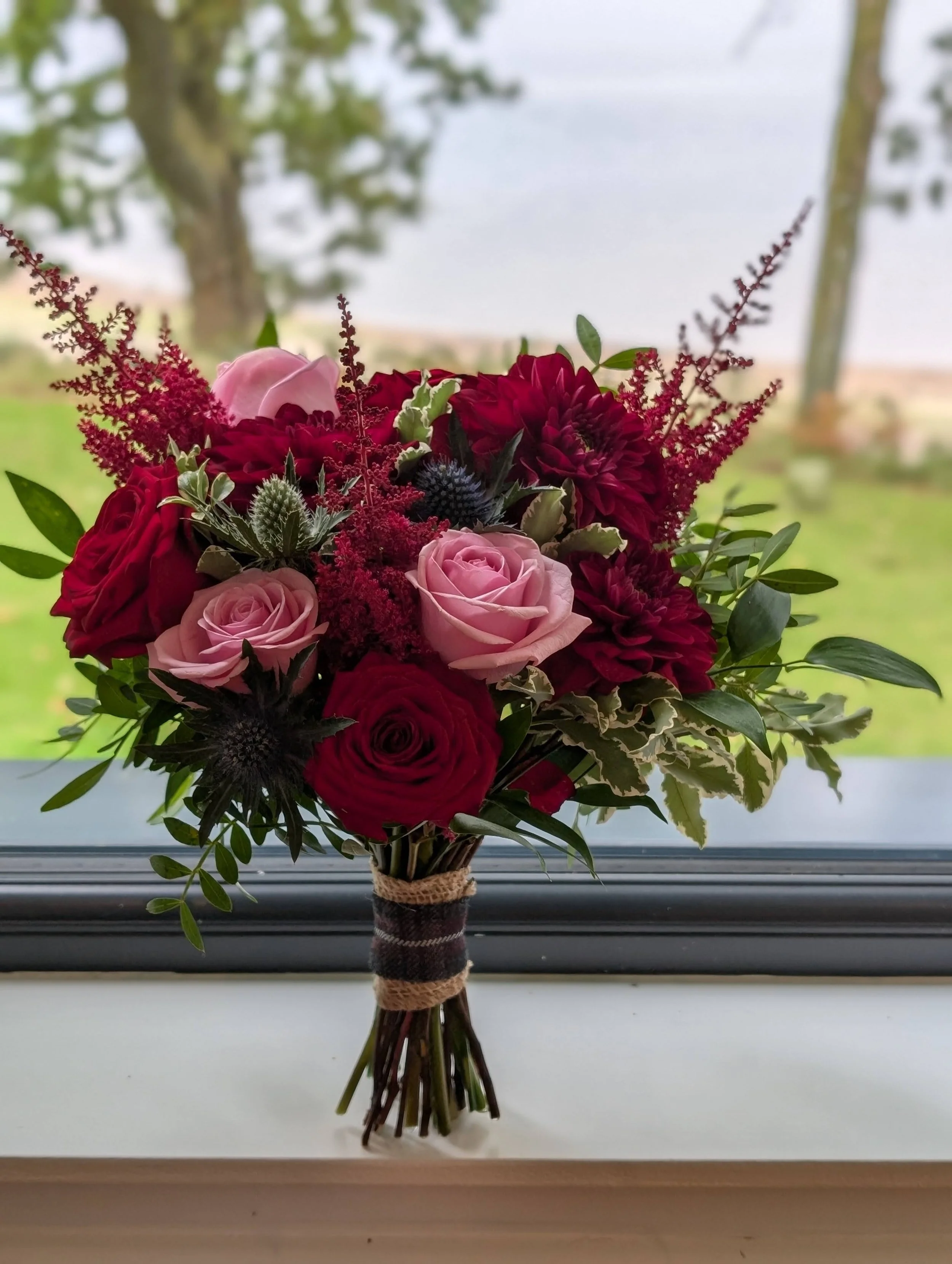 Bouquet of red and pink roses, dahlias, and greenery on a windowsill with a lake and trees in the background.