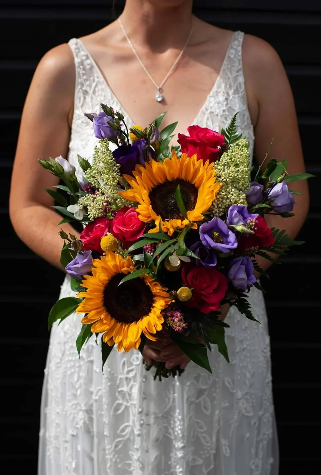 Person in a white lace dress holding a bouquet of sunflowers, roses, purple flowers, and greenery against a dark background.