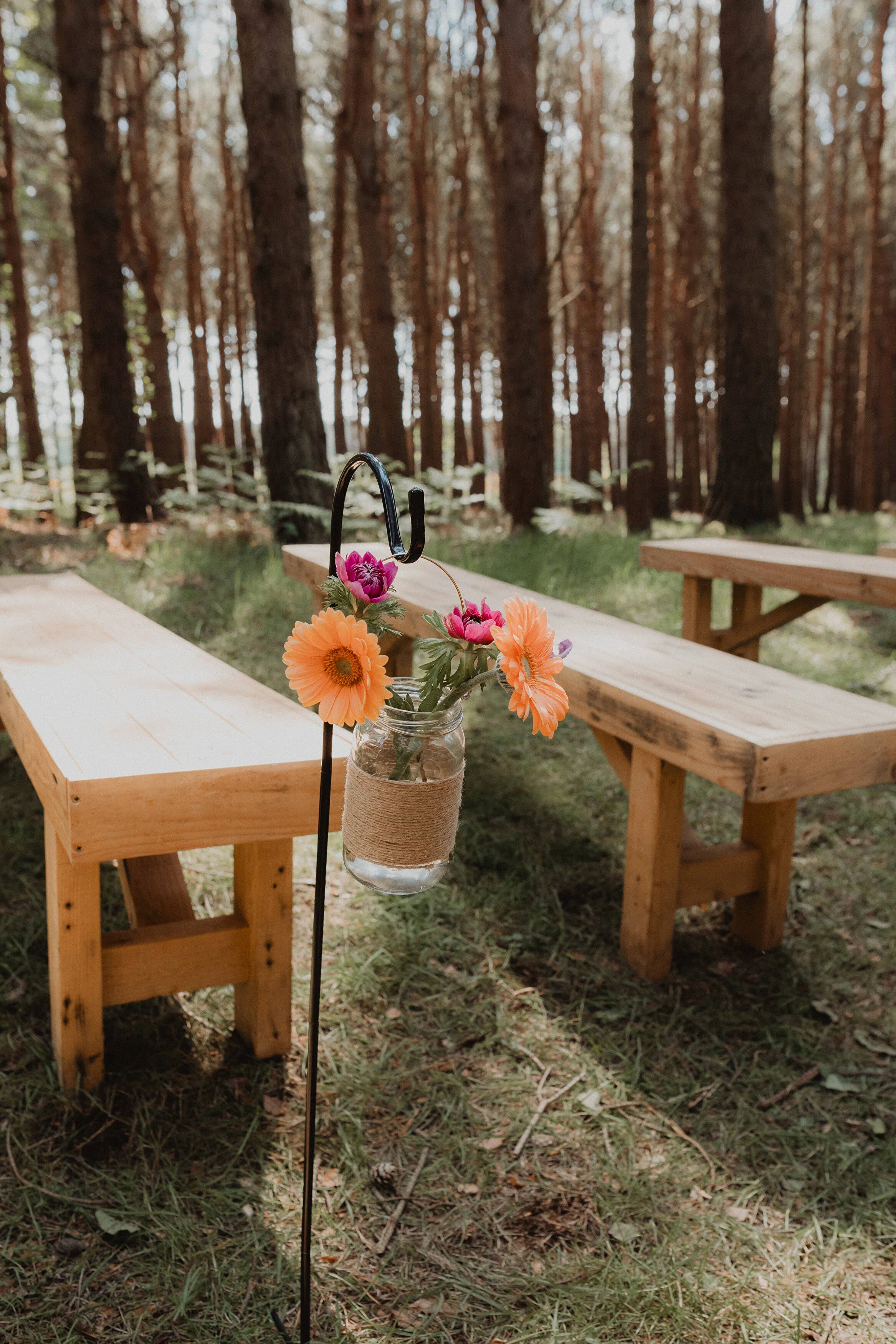 A rustic outdoor table setting with wooden benches in a forest, featuring a glass jar with pink and orange flowers hanging from a black metal stand.