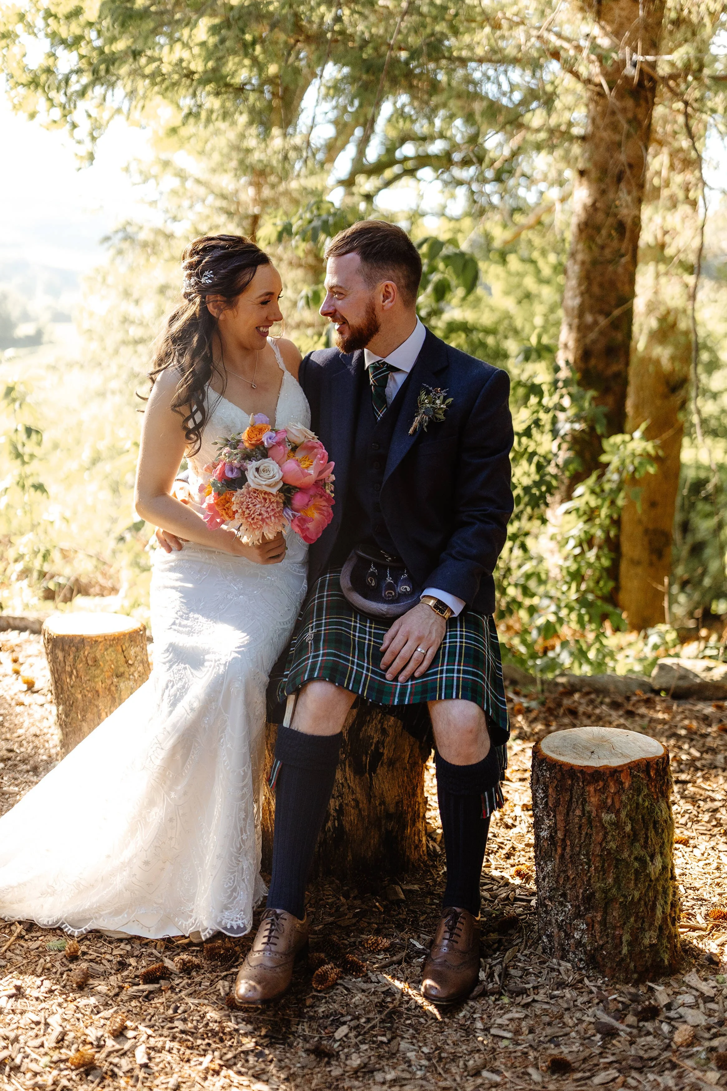 A bride and groom sitting on tree stumps in a forest, smiling and looking at each other. The bride is holding a bouquet of pink and white flowers, and the groom is dressed in traditional Scottish attire including a kilt, jacket, and tie.