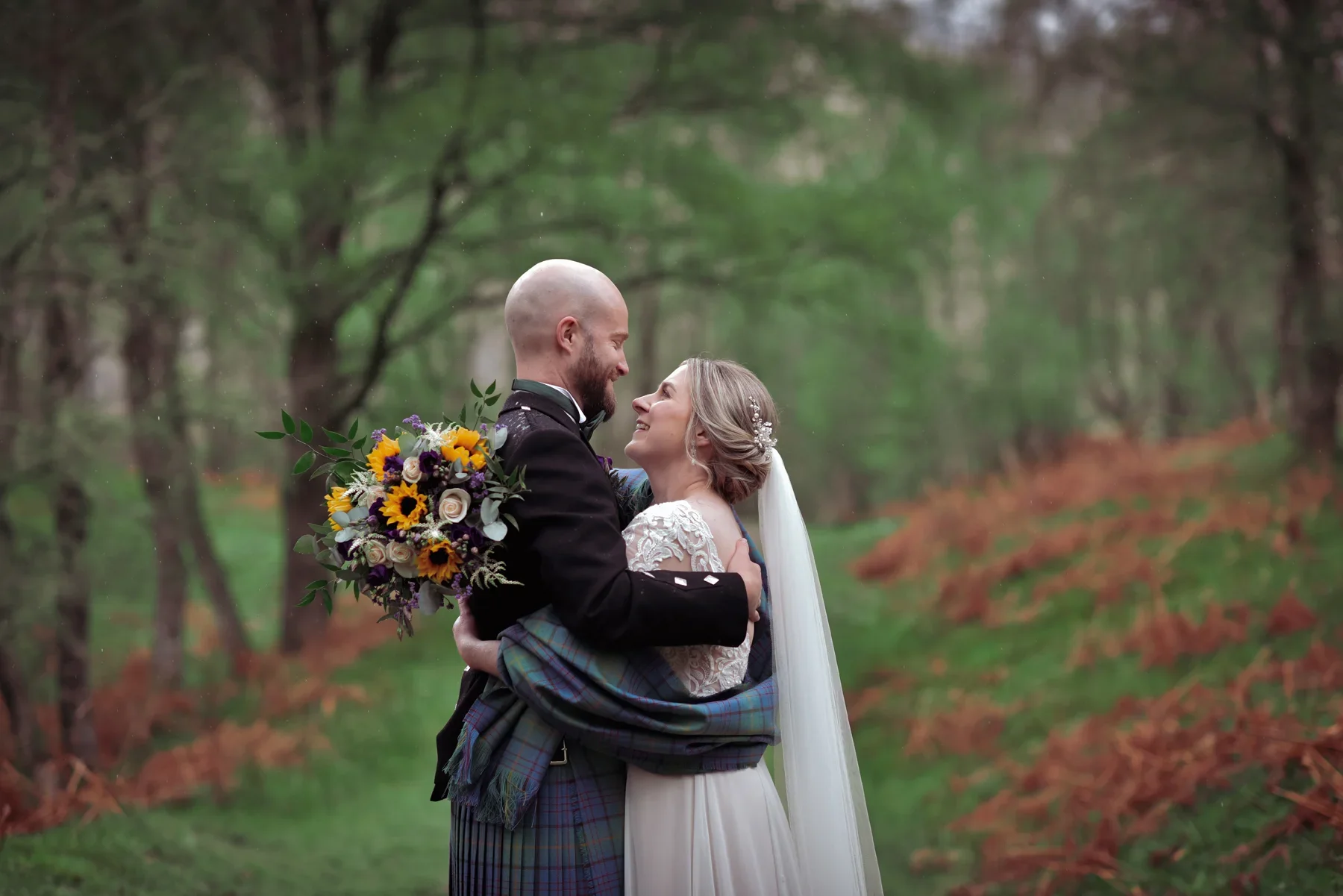 A bride and groom embracing outdoors in a wooded area, the groom in traditional Scottish attire holding a bouquet, and the bride in a lace wedding dress with a veil, smiling at each other.