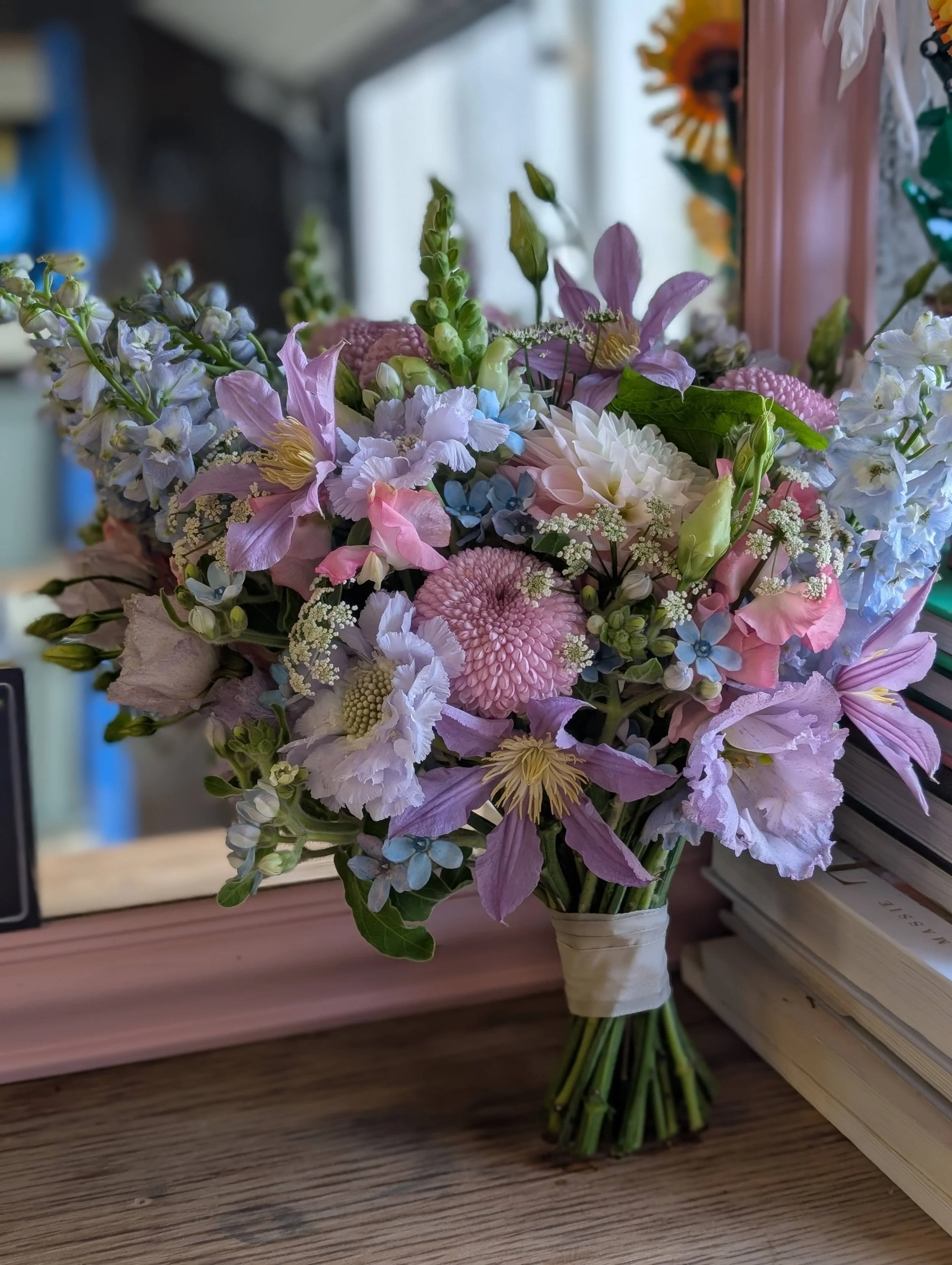 A colorful bouquet of flowers including pink, purple, white, and blue blooms, arranged on a wooden surface near a mirror.