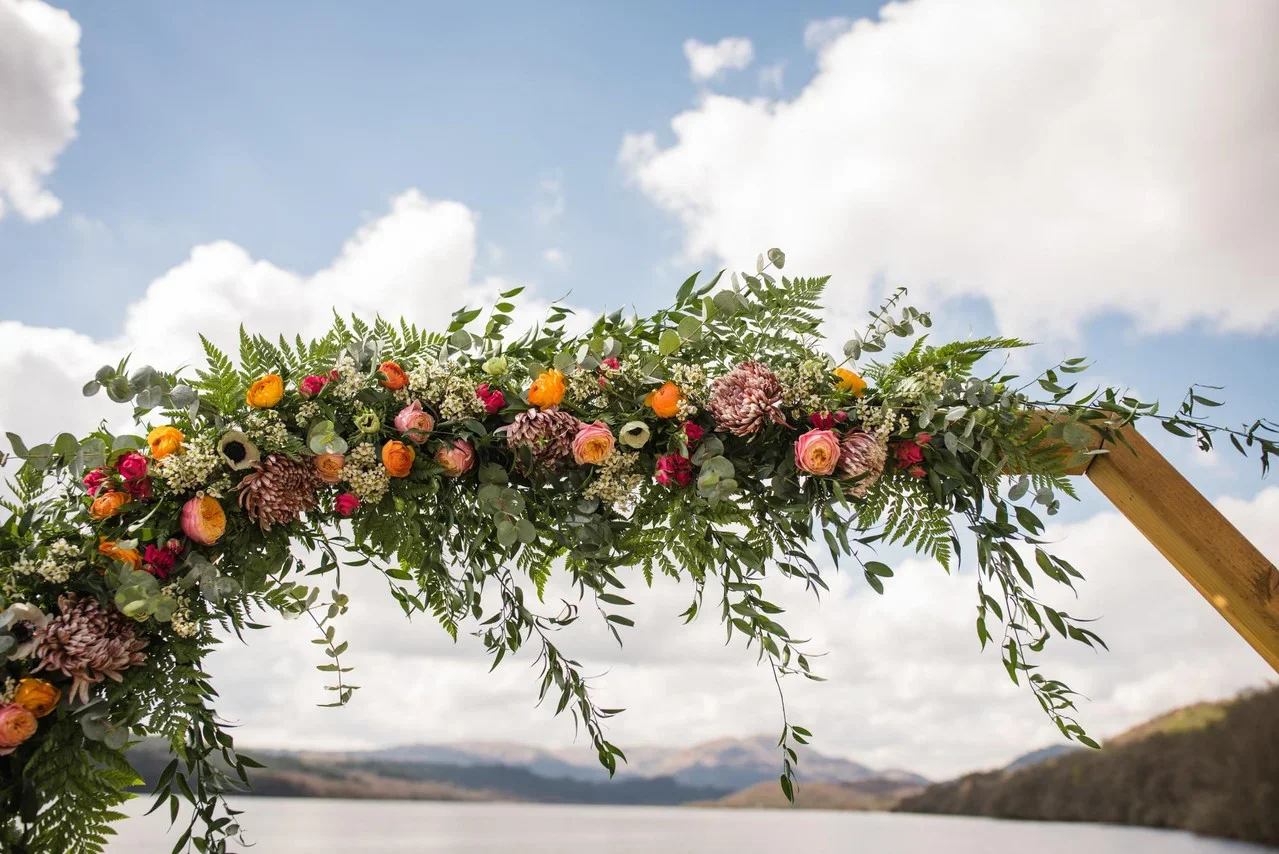 A floral arch with pink, yellow, and orange flowers and green foliage, overlooking a body of water and distant mountains under a cloudy sky.