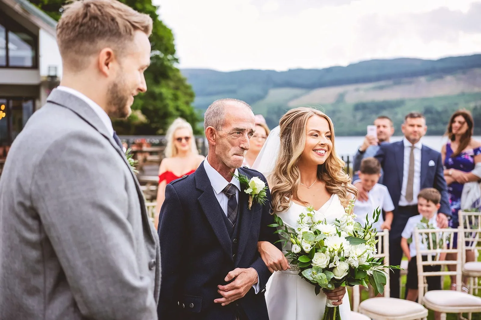 Bride smiling and holding a bouquet, walking down the aisle with her father at an outdoor wedding ceremony, guests seated and standing, scenic mountains in the background.