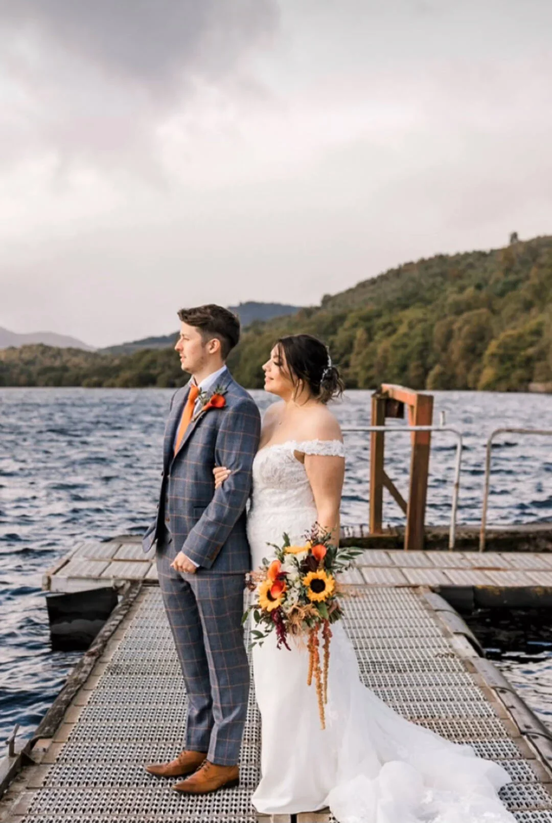 A bride and groom on a dock by a lake, posing for wedding photos with a scenic mountain background. The bride is holding a bouquet of sunflowers and orange flowers. The groom is dressed in a checked plaid suit with brown shoes.