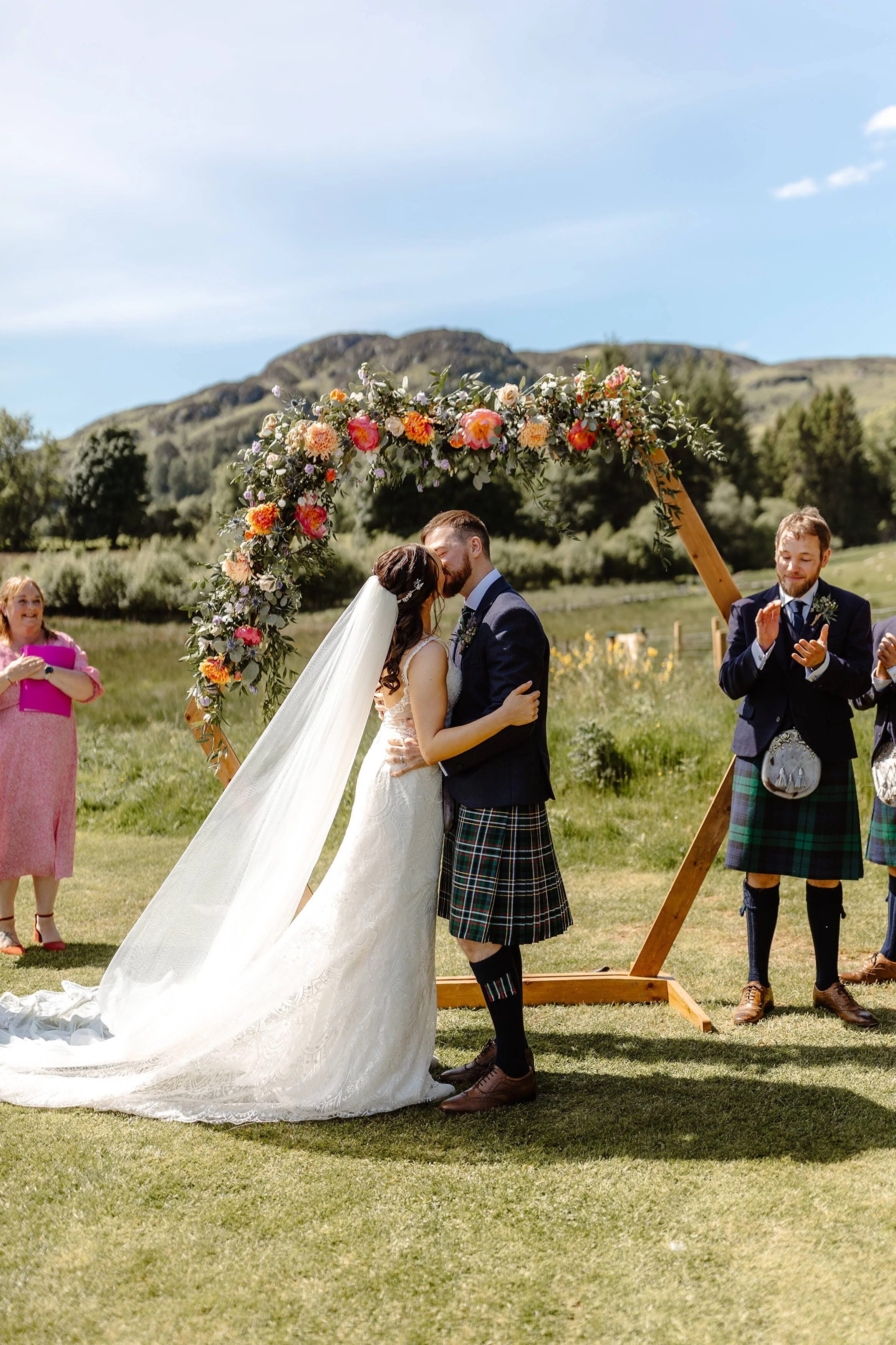 A couple in wedding attire kissing under a floral arch during an outdoor wedding ceremony with a scenic mountainous background.