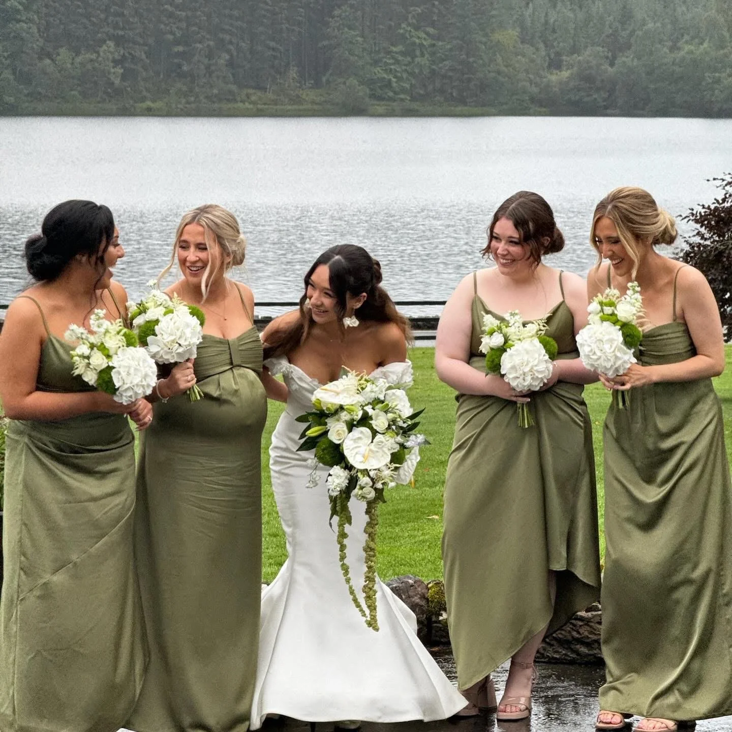 A group of five women dressed in wedding attire, standing outdoors near a body of water, smiling and holding bouquets of white flowers.
