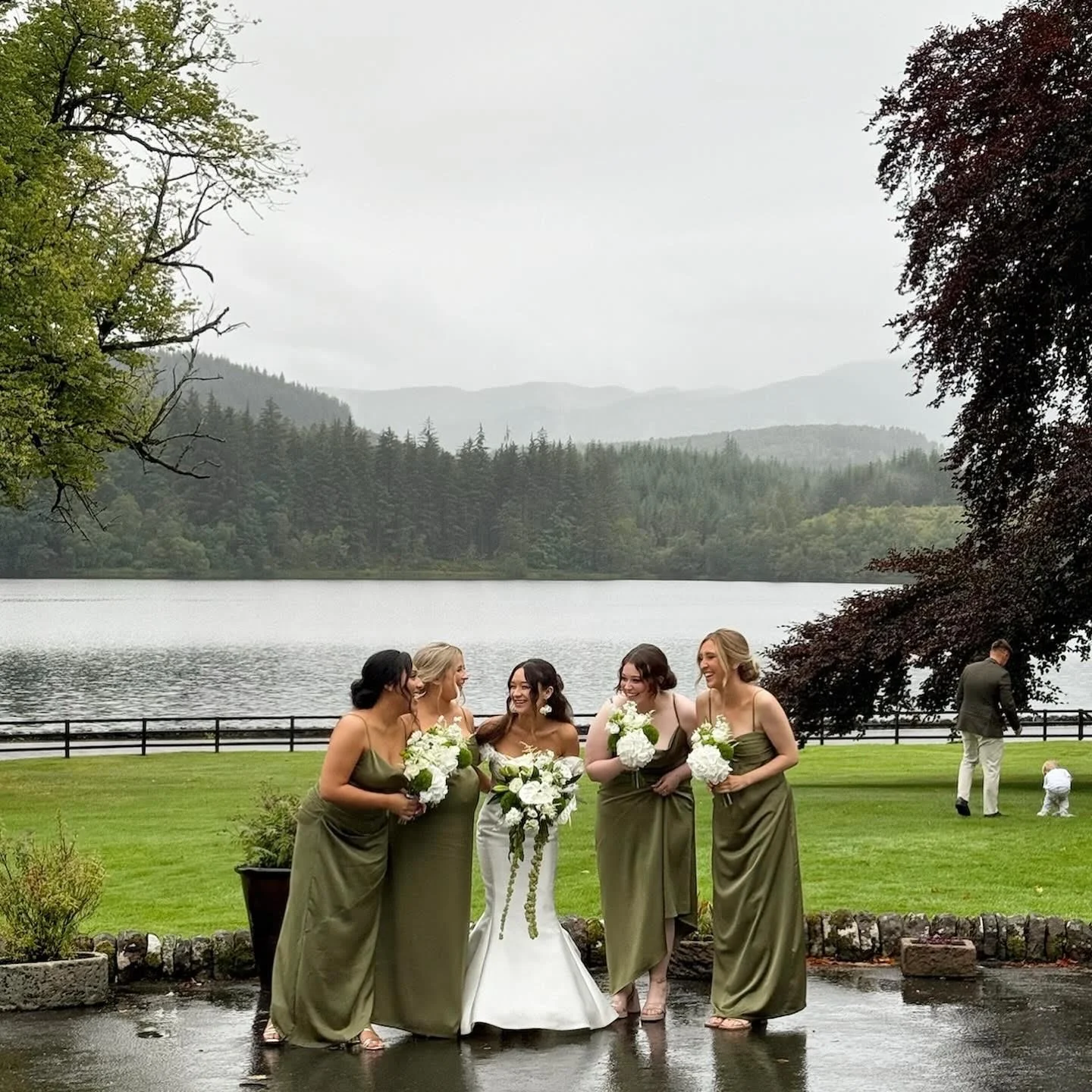 A bride and four bridesmaids standing together outdoors by a lake, holding bouquets of white flowers, with mountains and trees in the background on a cloudy day.