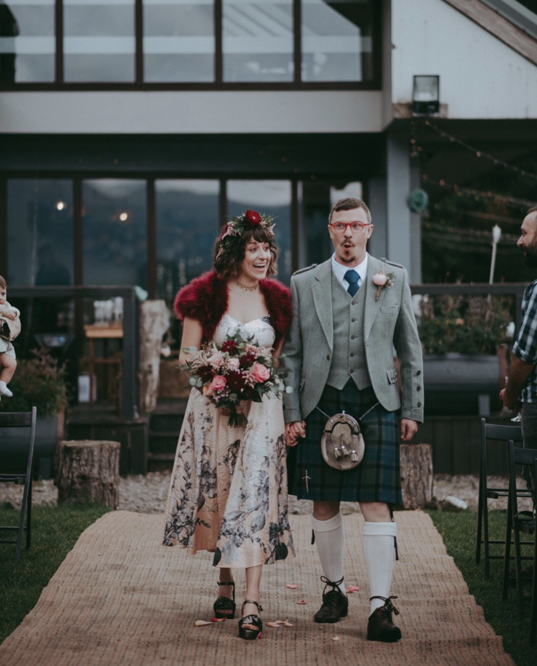 A couple holding hands walking down a beige aisle at their wedding, surrounded by friends and family, with a modern building in the background. The bride is dressed in a cream-colored floral dress with a red fur shawl and a flower crown, holding a bo