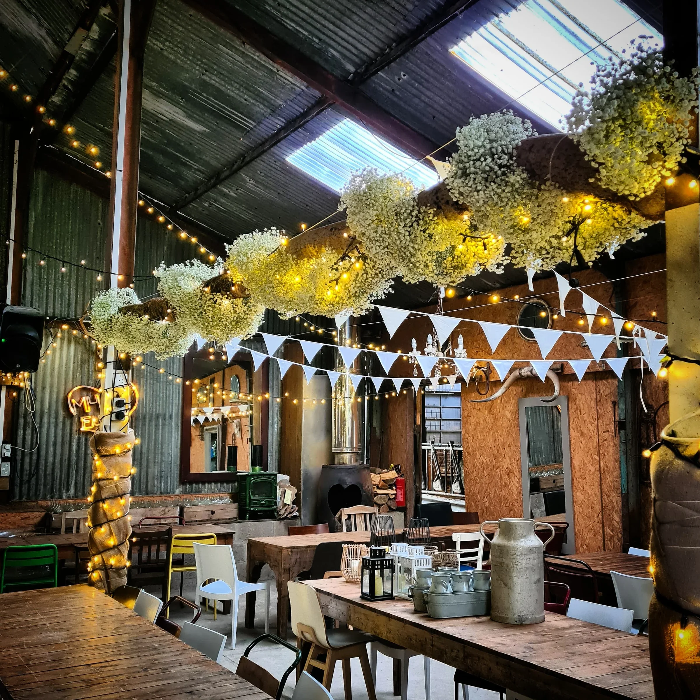 Decorated rustic indoor space with hanging white flowers, string lights, and white triangular pennant banners, featuring wooden tables and colorful chairs.