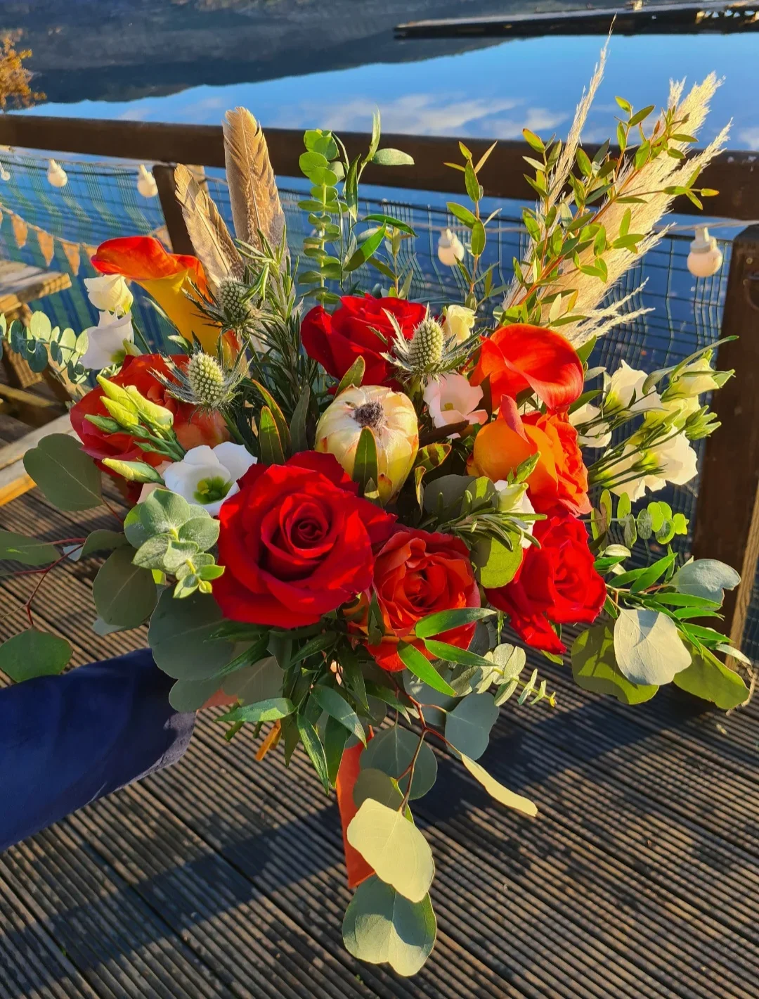 A colorful bouquet of flowers including red roses, white flowers, orange flowers, and greenery, held outdoors on a wooden deck near water with mountains in the background.
