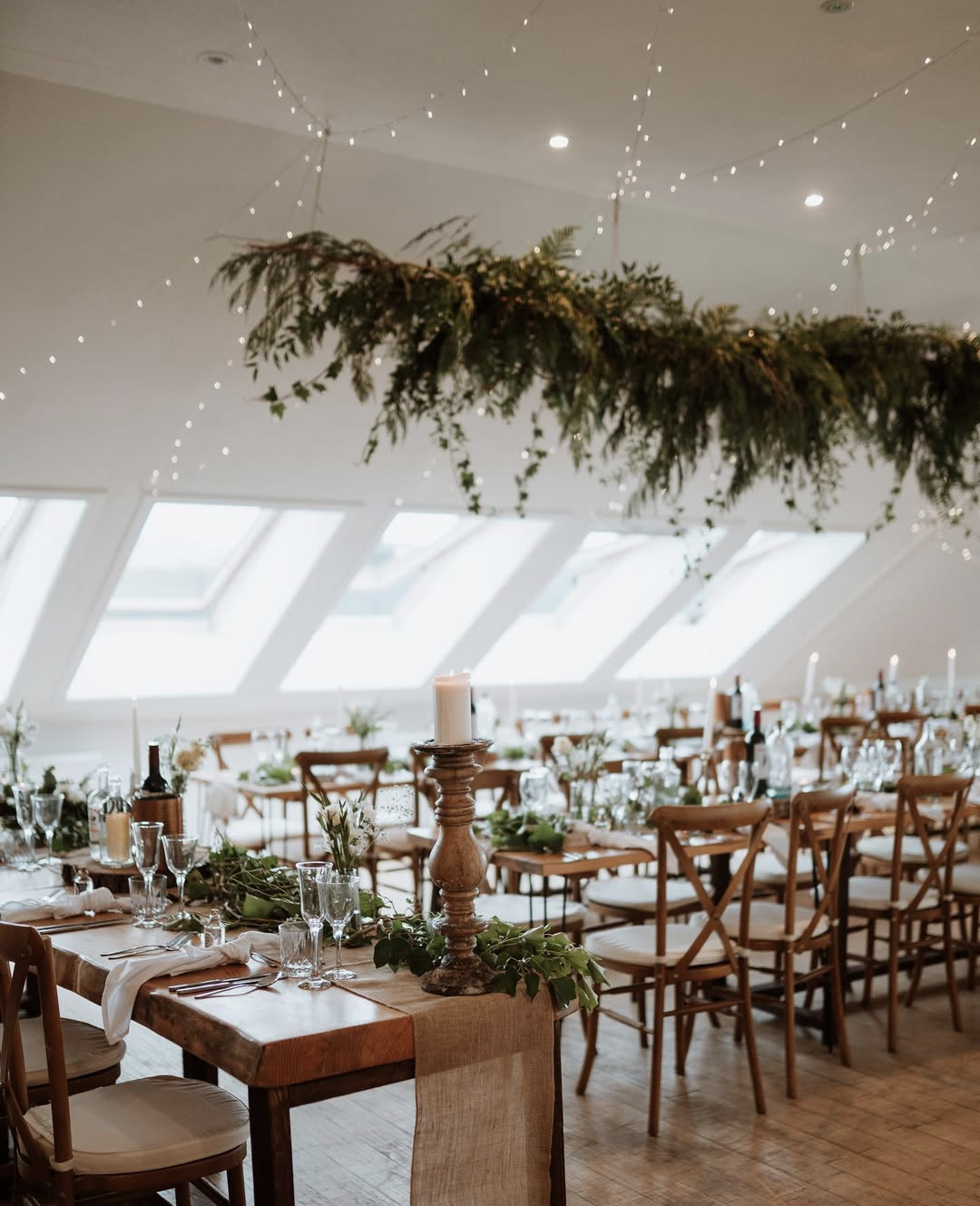 Wedding reception setup with wooden tables decorated with greenery, white flowers, and candles inside a bright room with ceiling string lights and skylights.