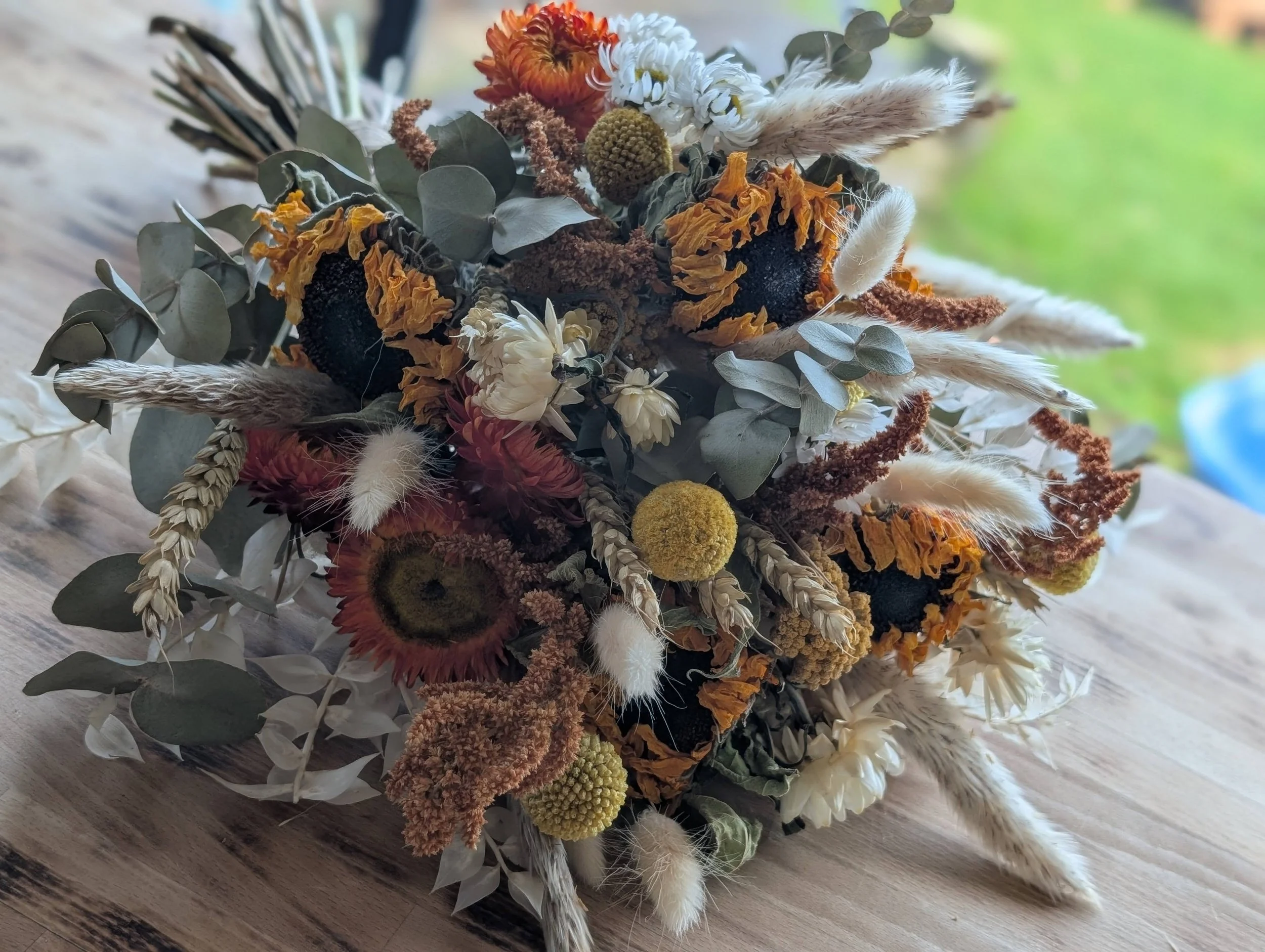 Dried floral arrangement featuring sunflowers, eucalyptus leaves, white and cream flowers, and various dried grasses and seed pods, placed on a wooden surface with a blurred green outdoor background.