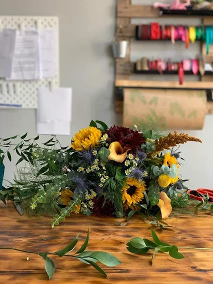 A colorful flower arrangement featuring sunflowers, calla lilies, and various greenery on a wooden table, with a workshop or craft room in the background.