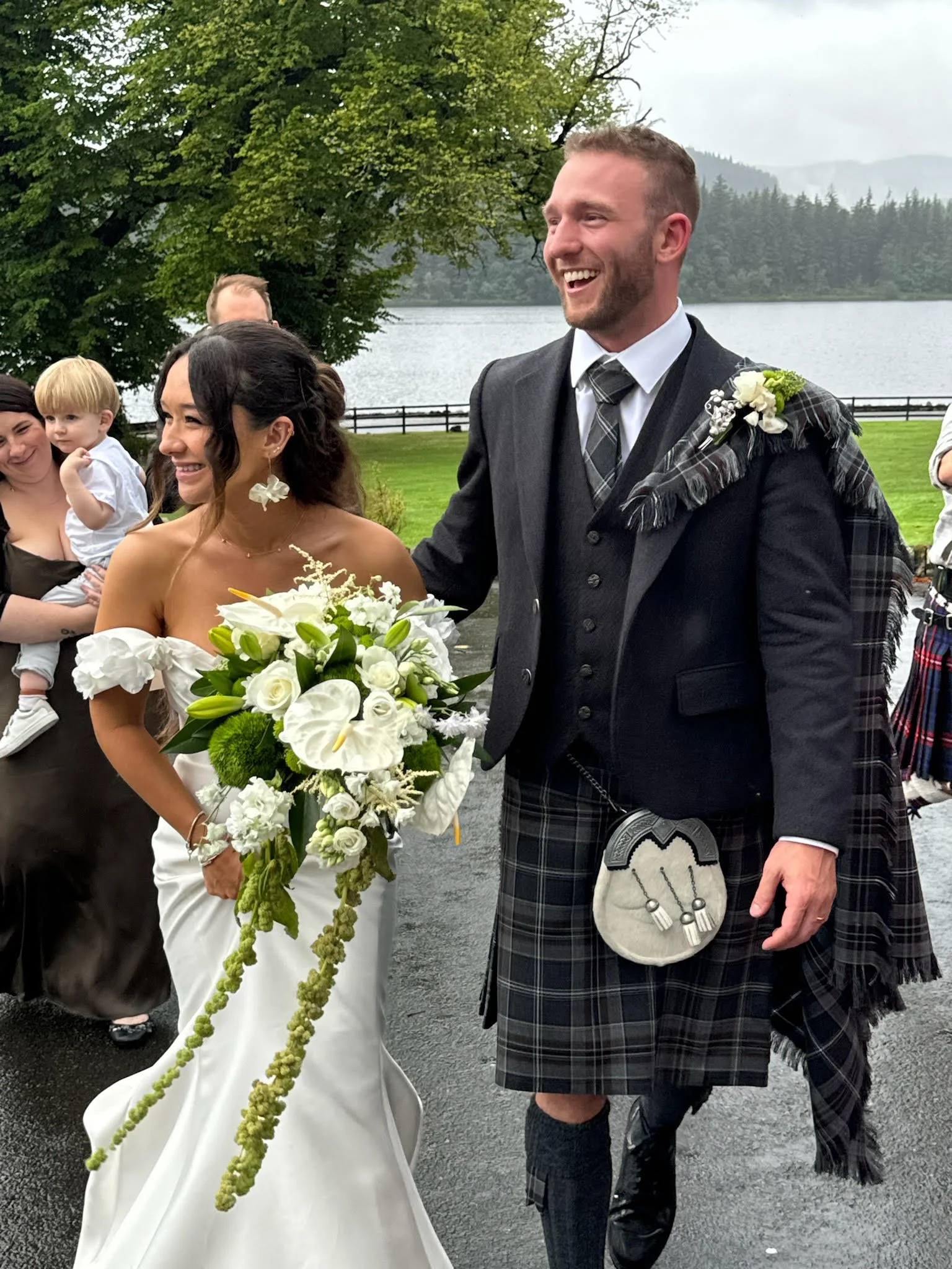 A happy bride and groom at their wedding outdoors by a lake, with guests in the background. The bride is holding a large white floral bouquet and wearing a strapless white wedding dress. The groom is dressed in traditional Scottish attire, including 