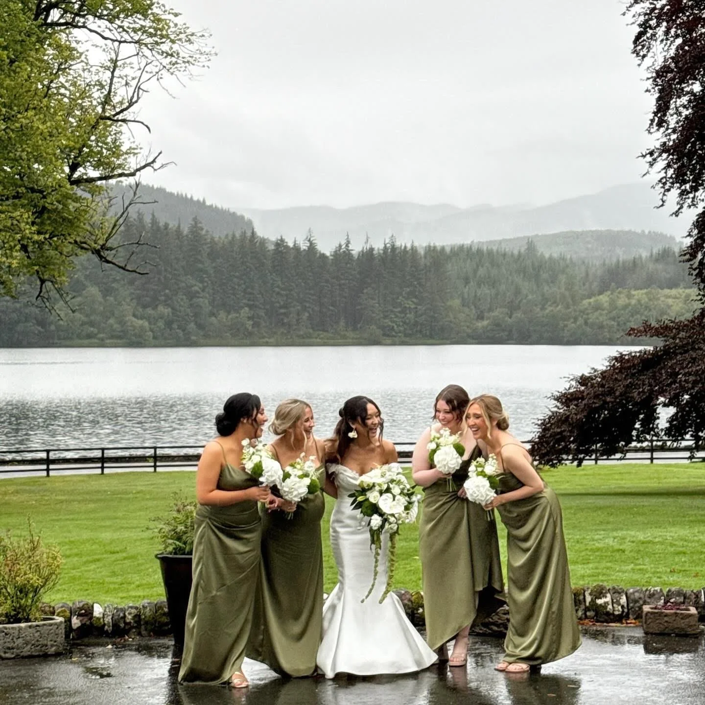 Bride and five bridesmaids standing together outdoors near a lake with trees and mountains in the background, holding bouquets of white flowers.