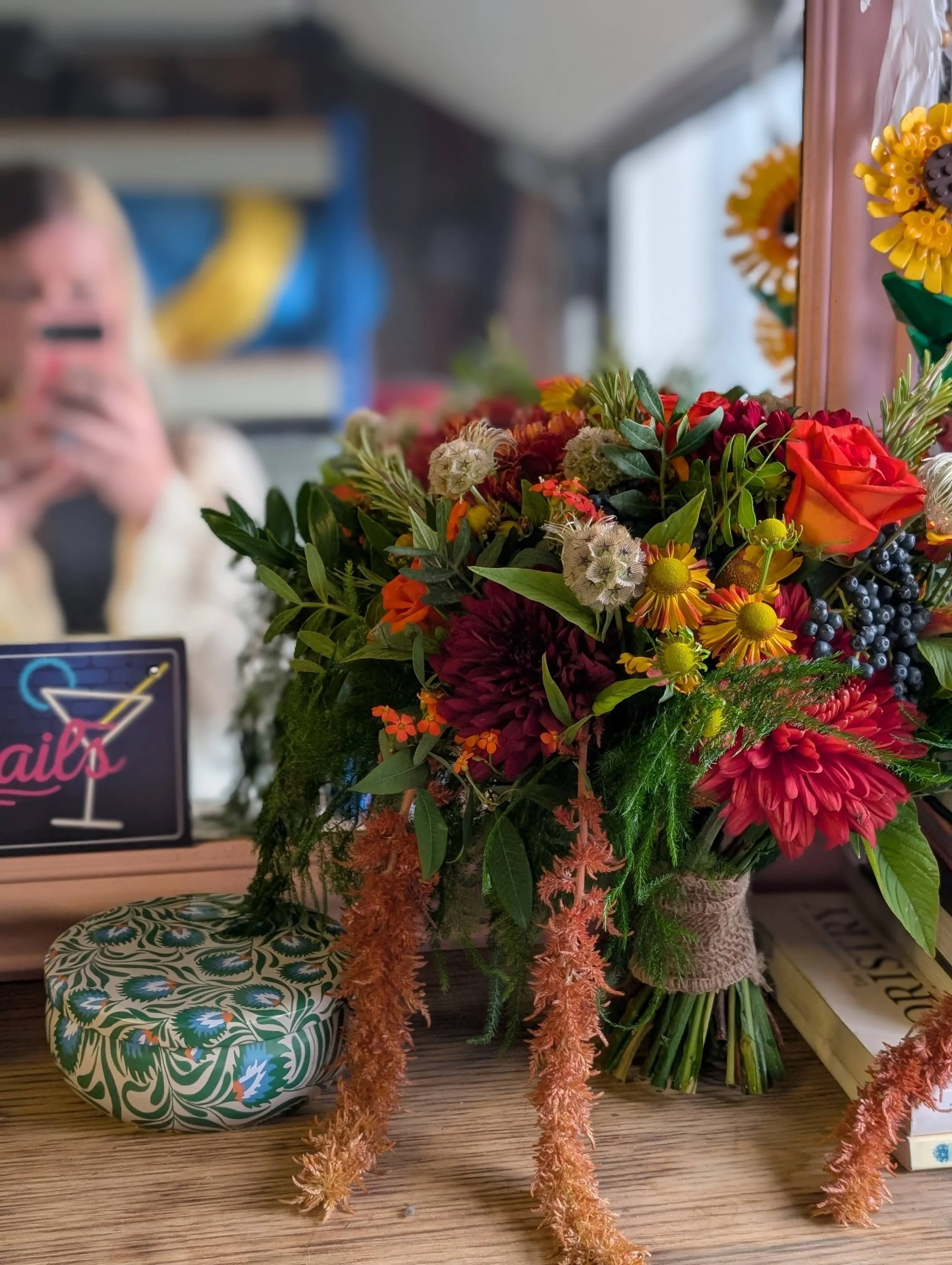 A colorful bouquet of flowers including roses, dahlias, and other blooms on a wooden table with a mirror, a decorative box, and a neon sign that reads 'nails' in the background.
