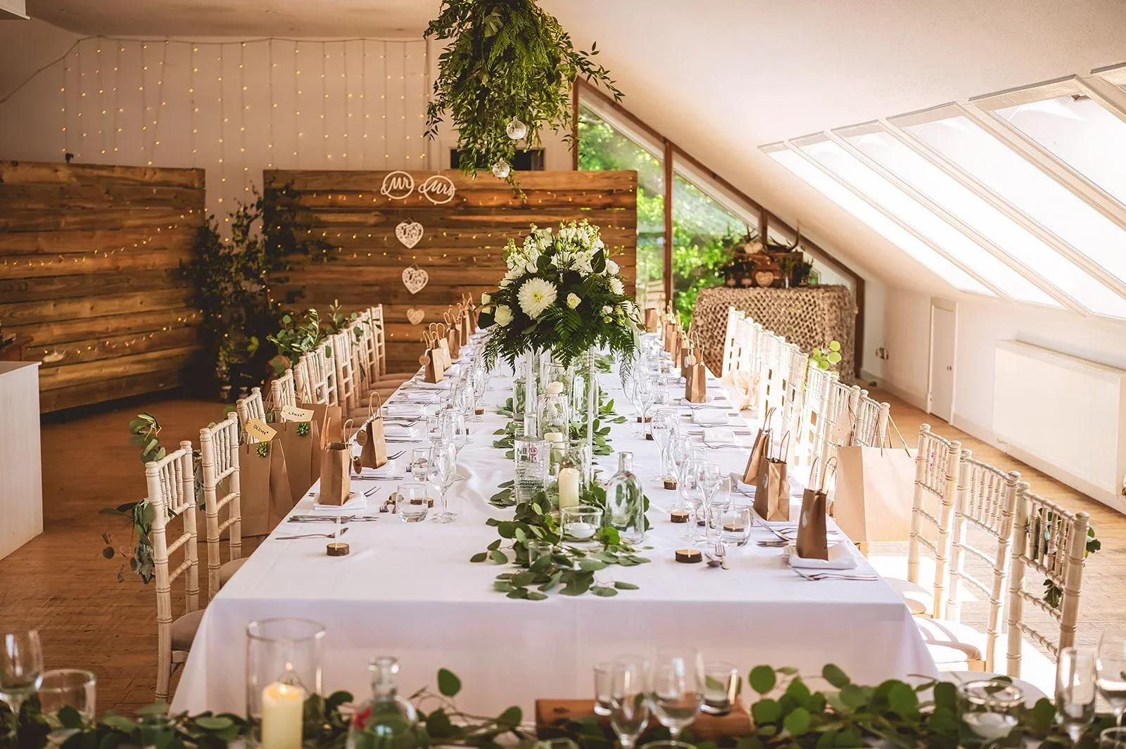 Wedding reception table with white tablecloth, a long floral centerpiece with white flowers and greenery, surrounded by cream chairs decorated with greenery. The background features a wooden wall with string lights and hanging heart decorations, with